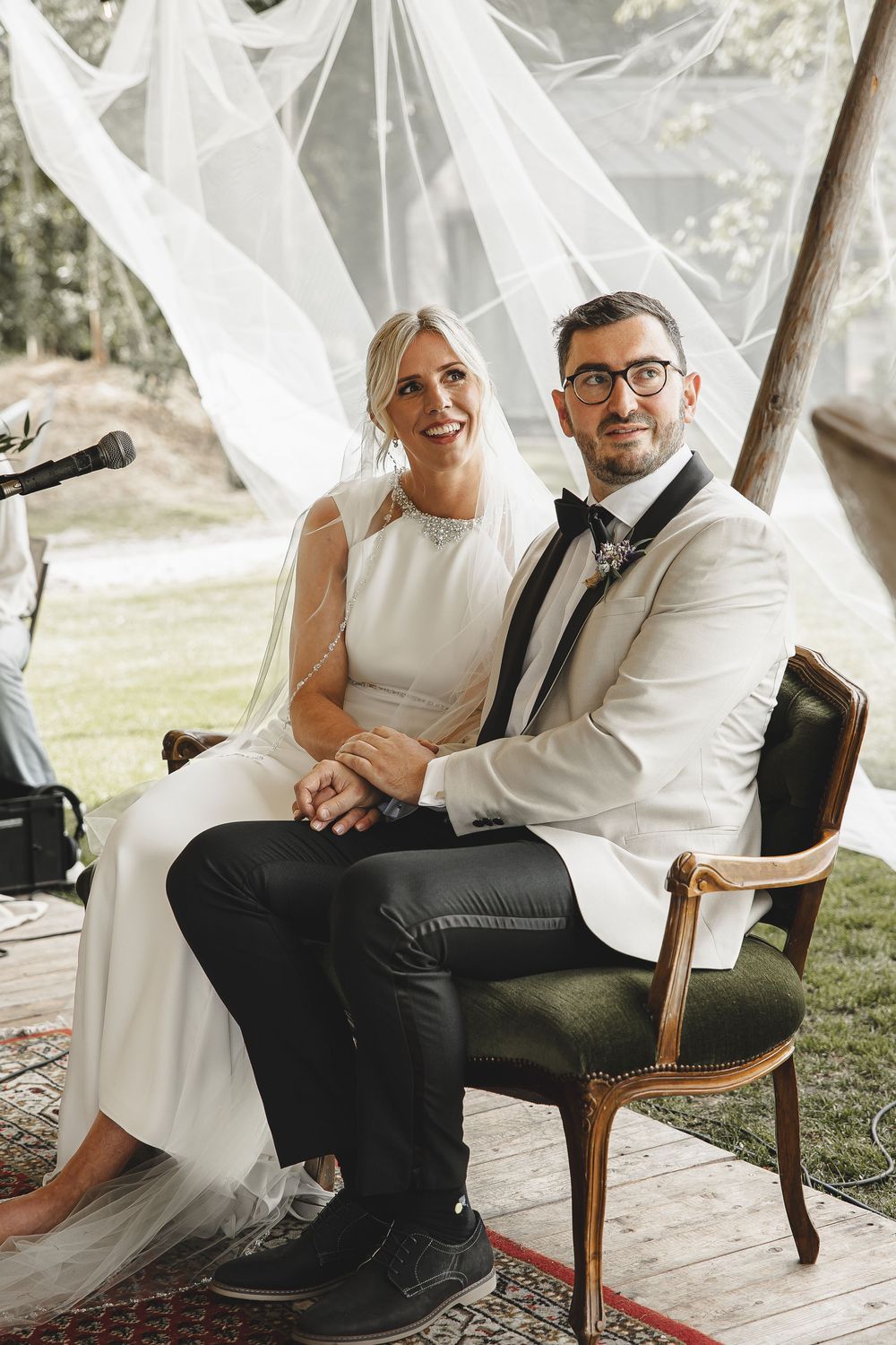 A bride and groom share a joyful moment while seated on a vintage chair under flowing white fabric at an outdoor wedding.