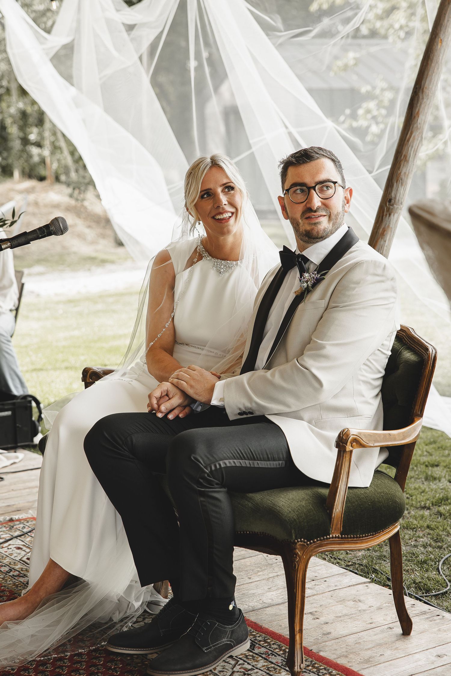 A bride and groom share a joyful moment while seated on a vintage chair under flowing white fabric at an outdoor wedding.