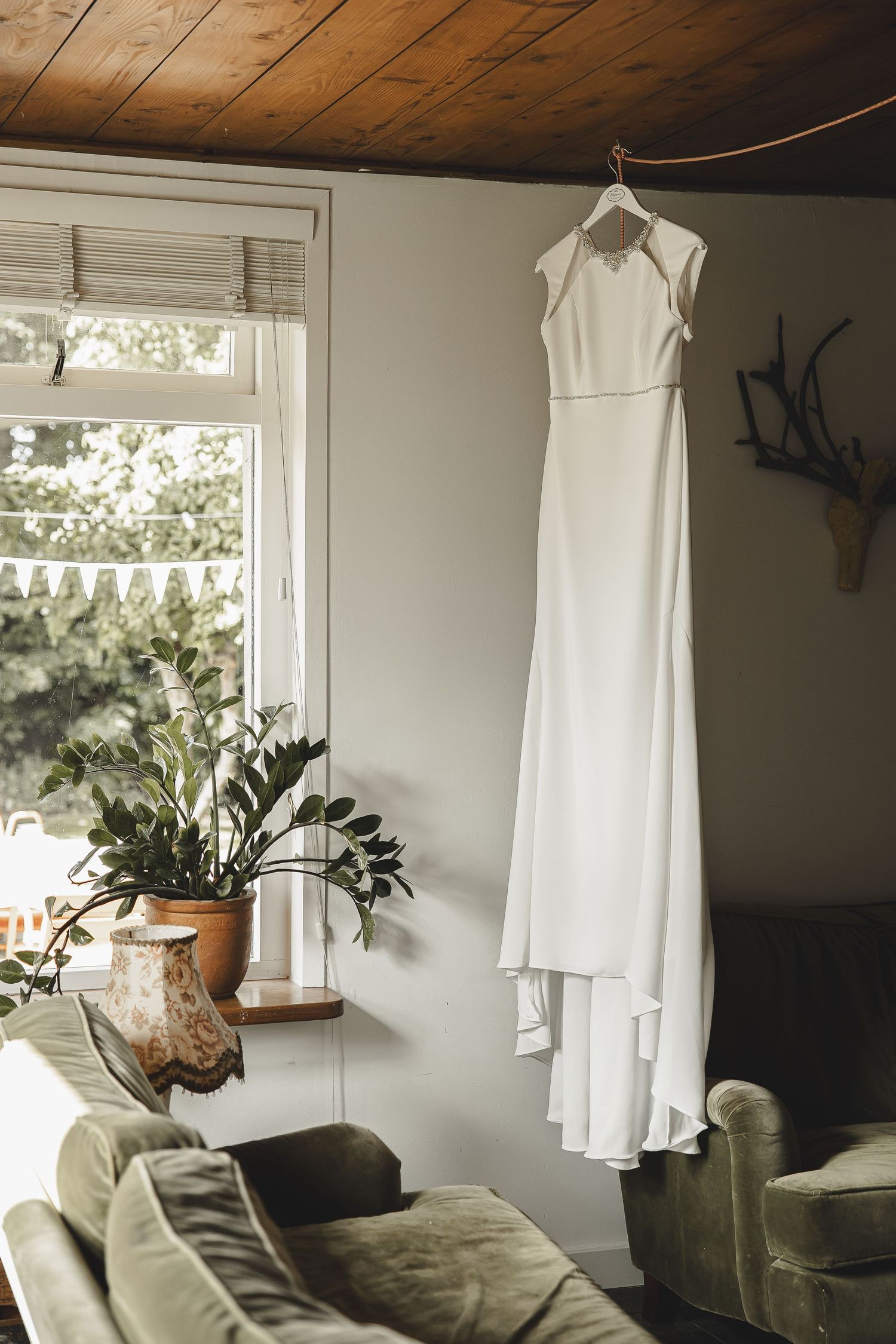 A white dress hangs near a window in a cozy room with olive green furniture and natural light.