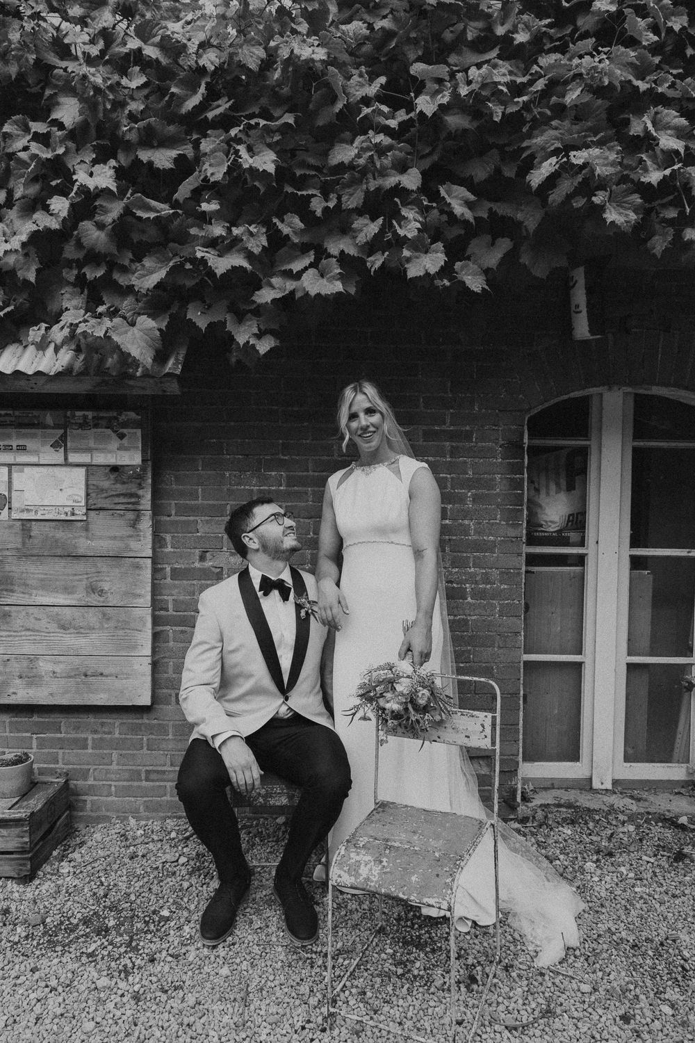 Black and white wedding photos of a couple sitting together on a grassy area near a brick building.