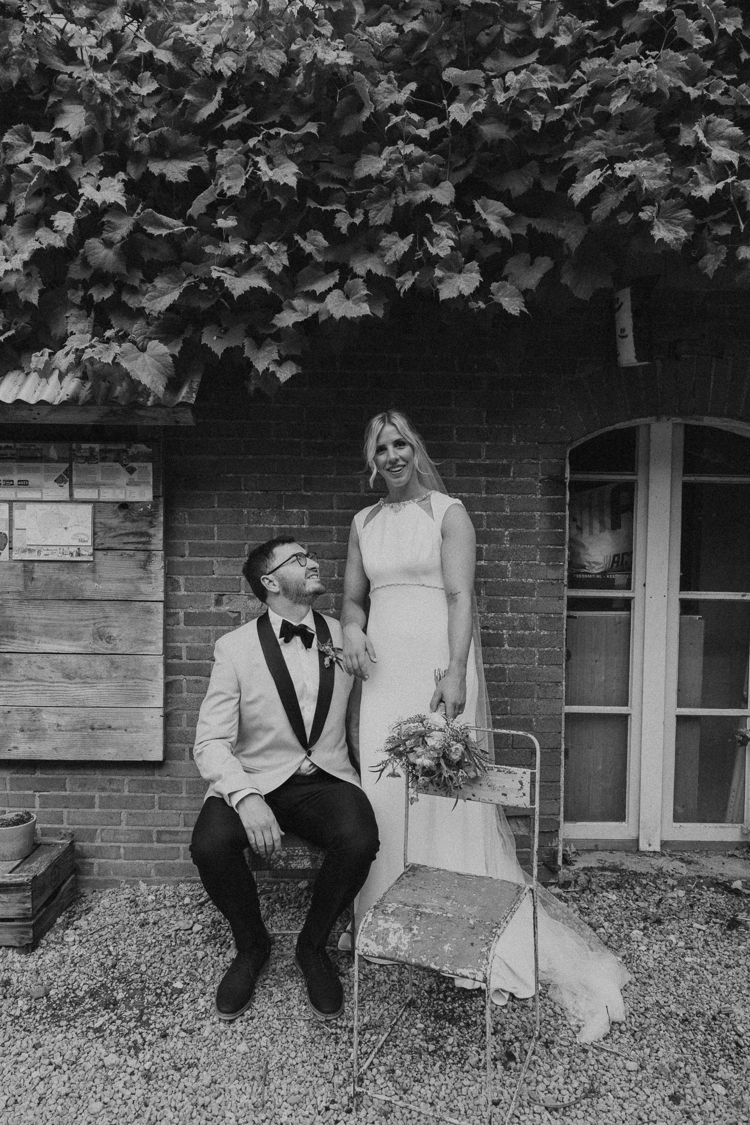 Black and white wedding photos of a couple sitting together on a grassy area near a brick building.