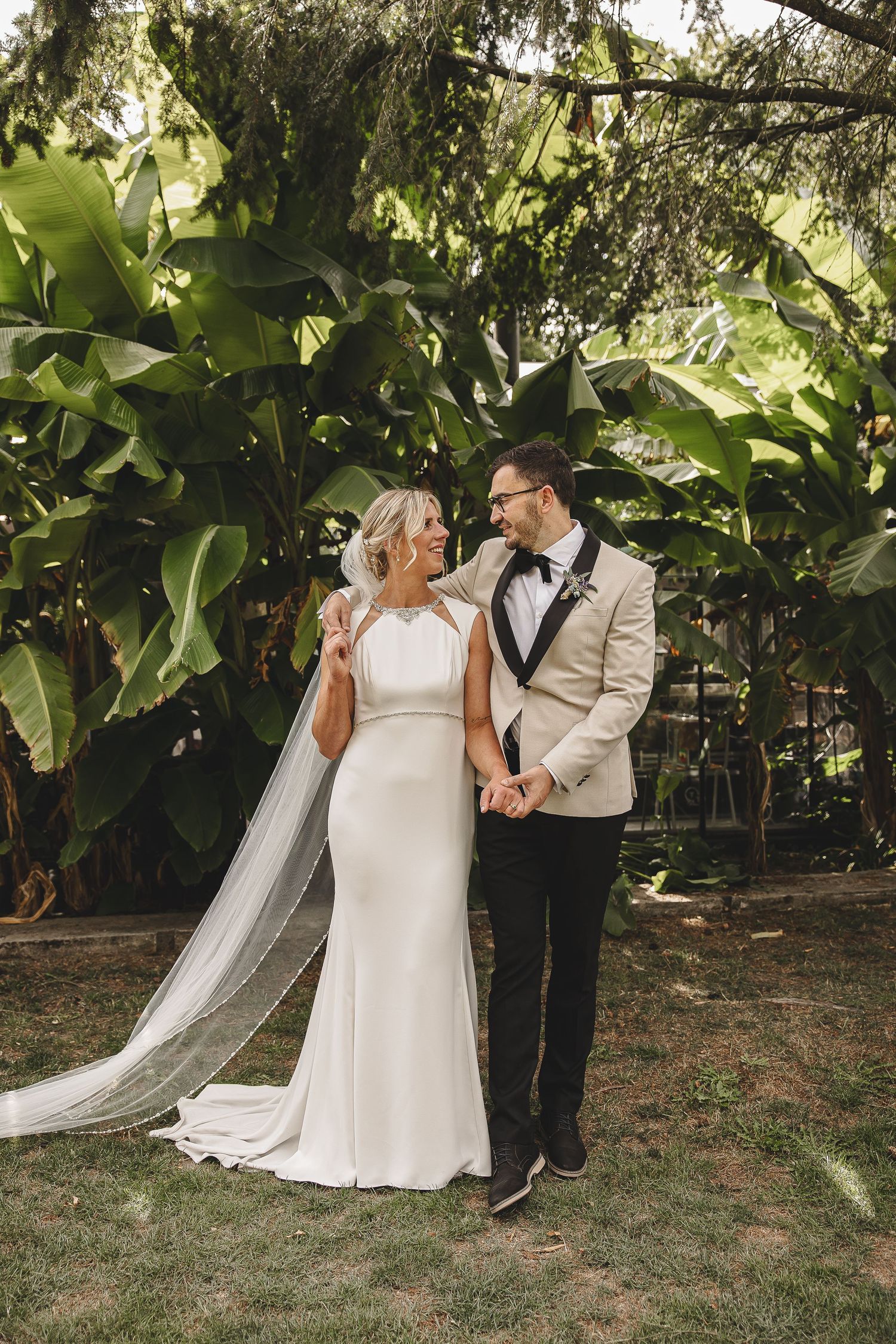 Newlywed couple shares romantic moments under lush green trees during outdoor garden wedding photoshoot.