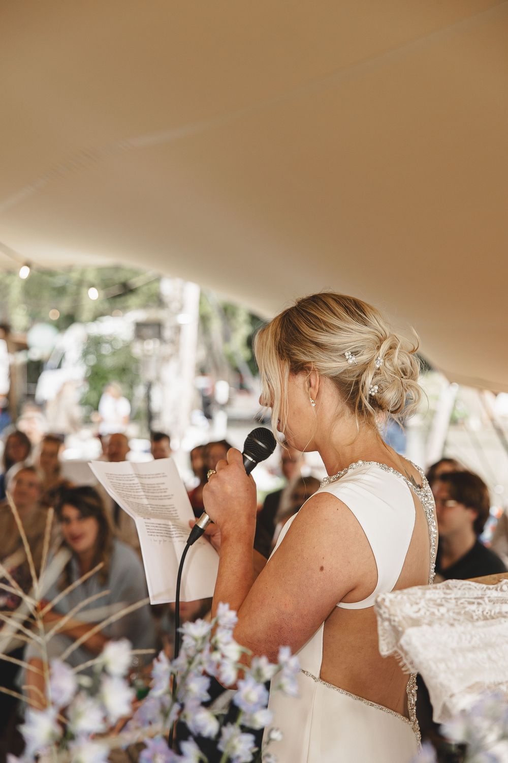 Bride speaking into microphone during outdoor wedding reception under tent with guests in background.