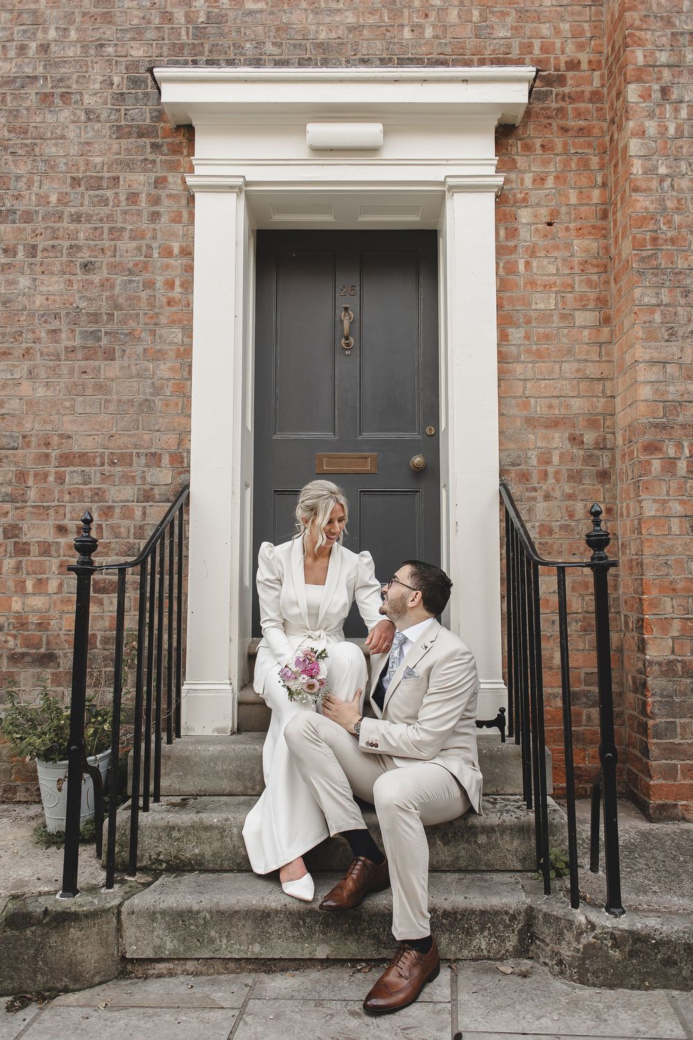Wedding couple poses playfully on brick building steps with classic white doorway.