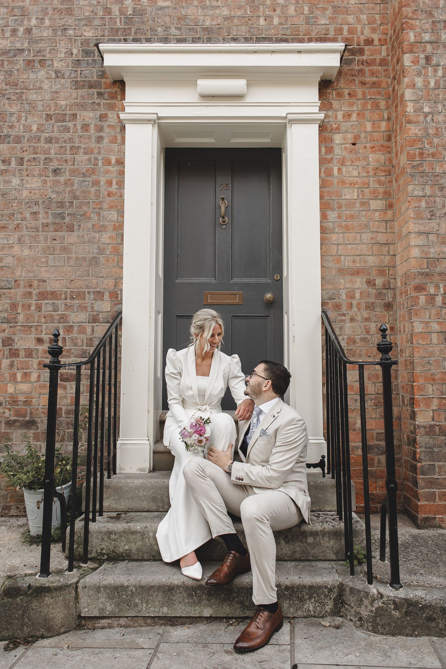 Wedding couple poses playfully on brick building steps with classic white doorway.