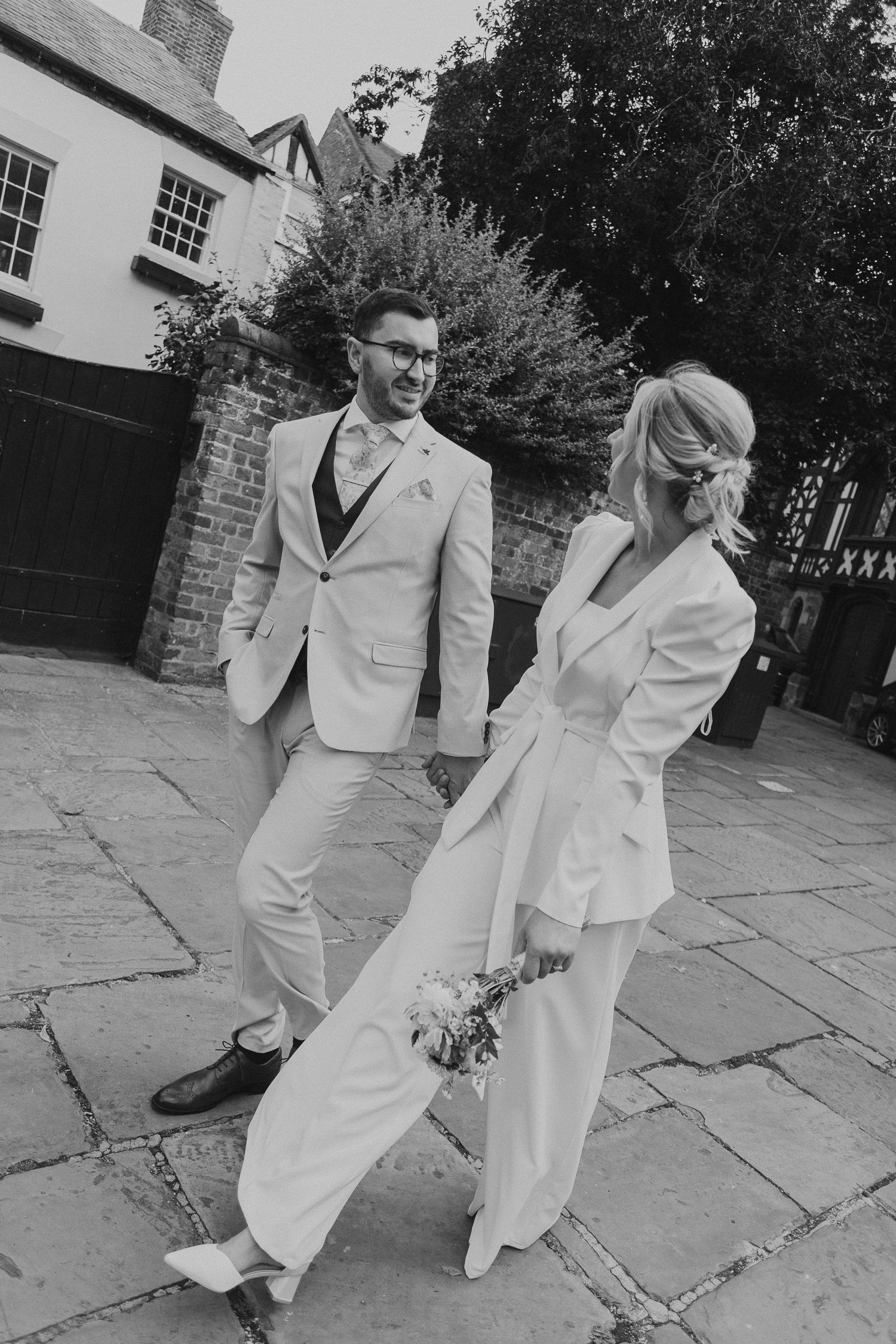 A couple in vintage white suits dances playfully on stone pavement in a black and white photograph.