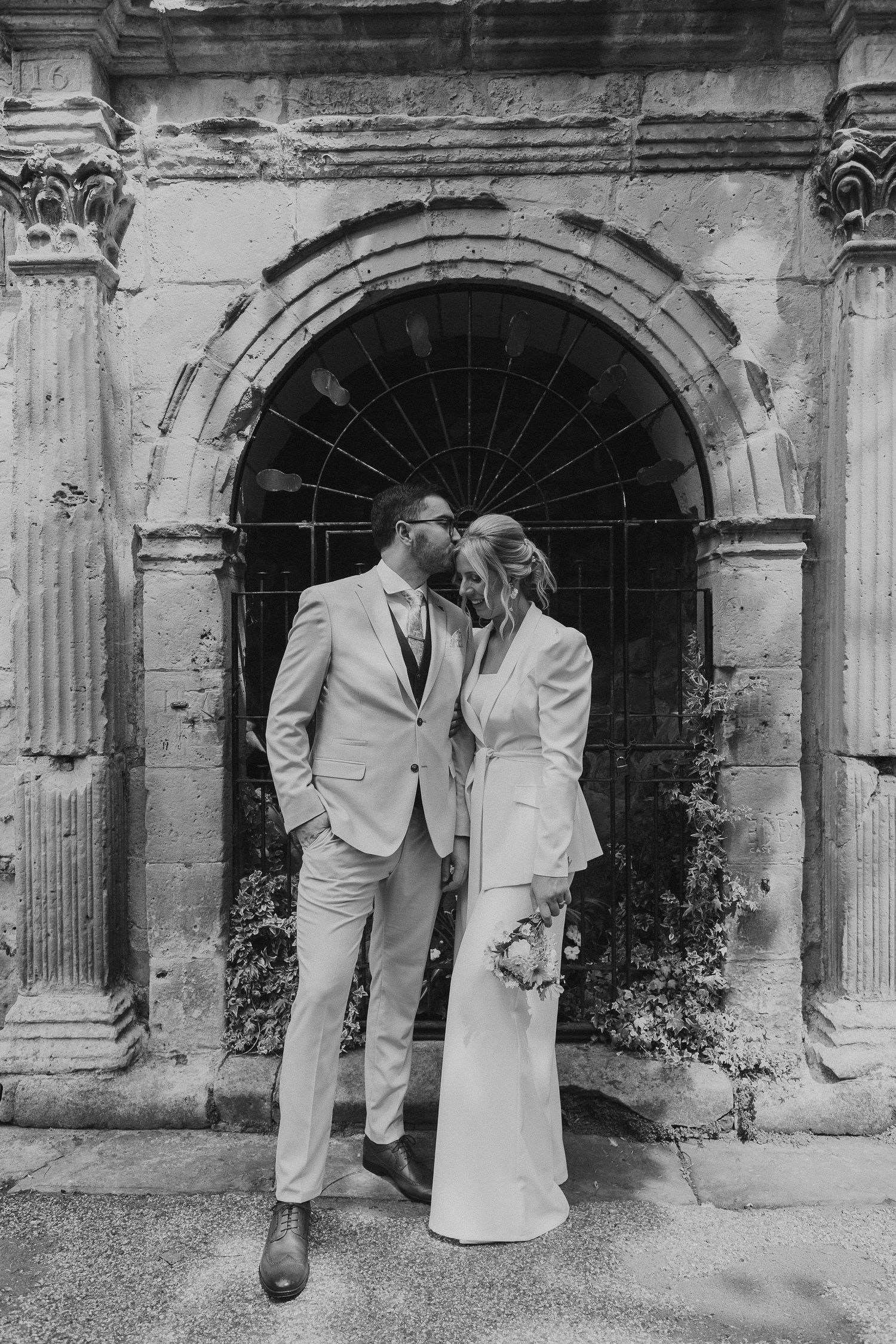 A couple in matching light-colored suits share an intimate moment in front of an ornate stone archway entrance.