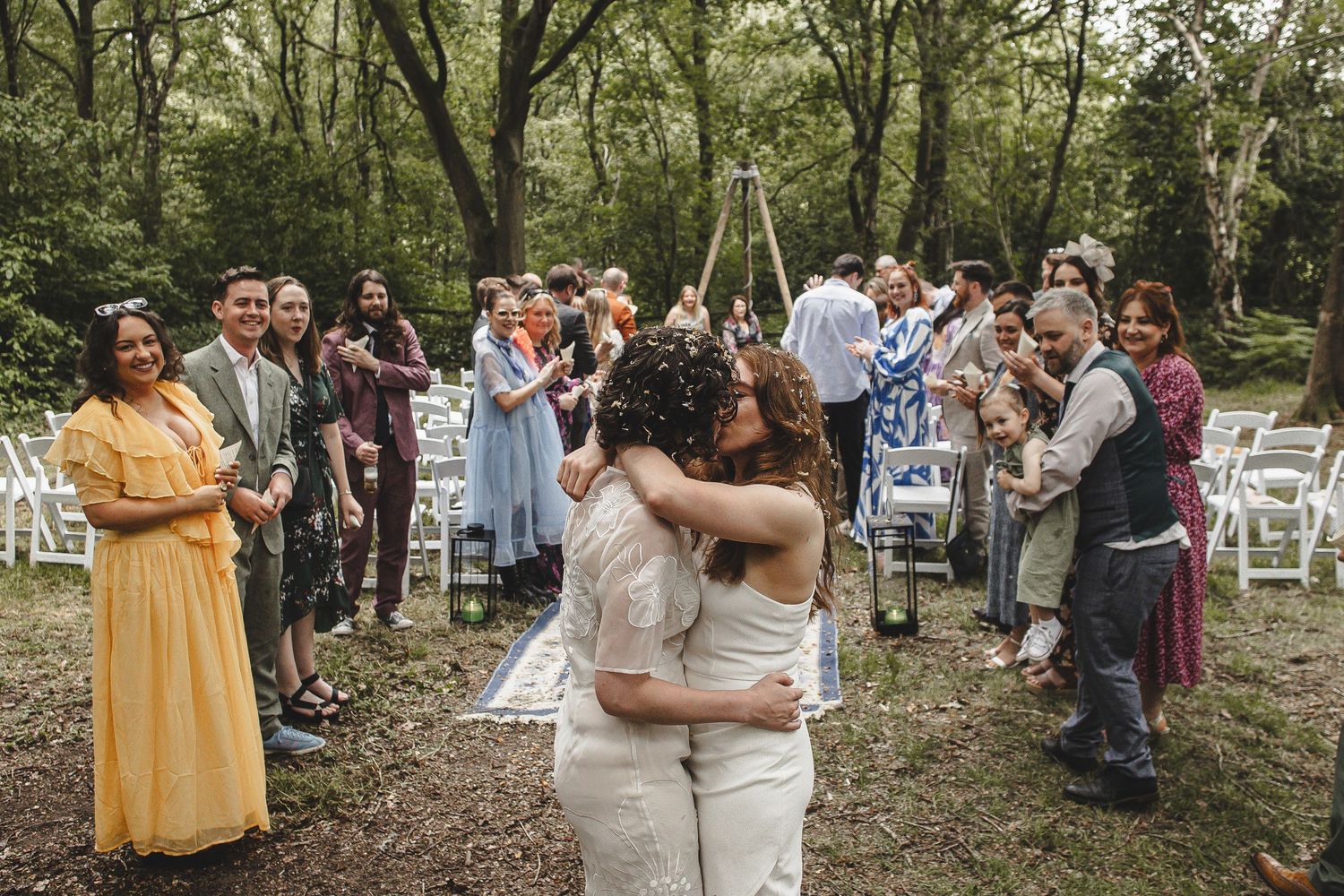 Two people embrace during an outdoor woodland wedding ceremony as guests look on from white chairs in a forest setting.
