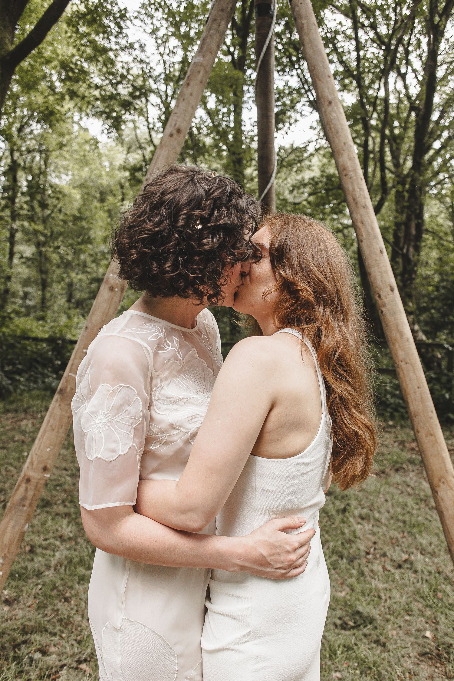 A romantic couple in white outfits share a tender kiss while embracing under wooden poles in a wooded forest setting.