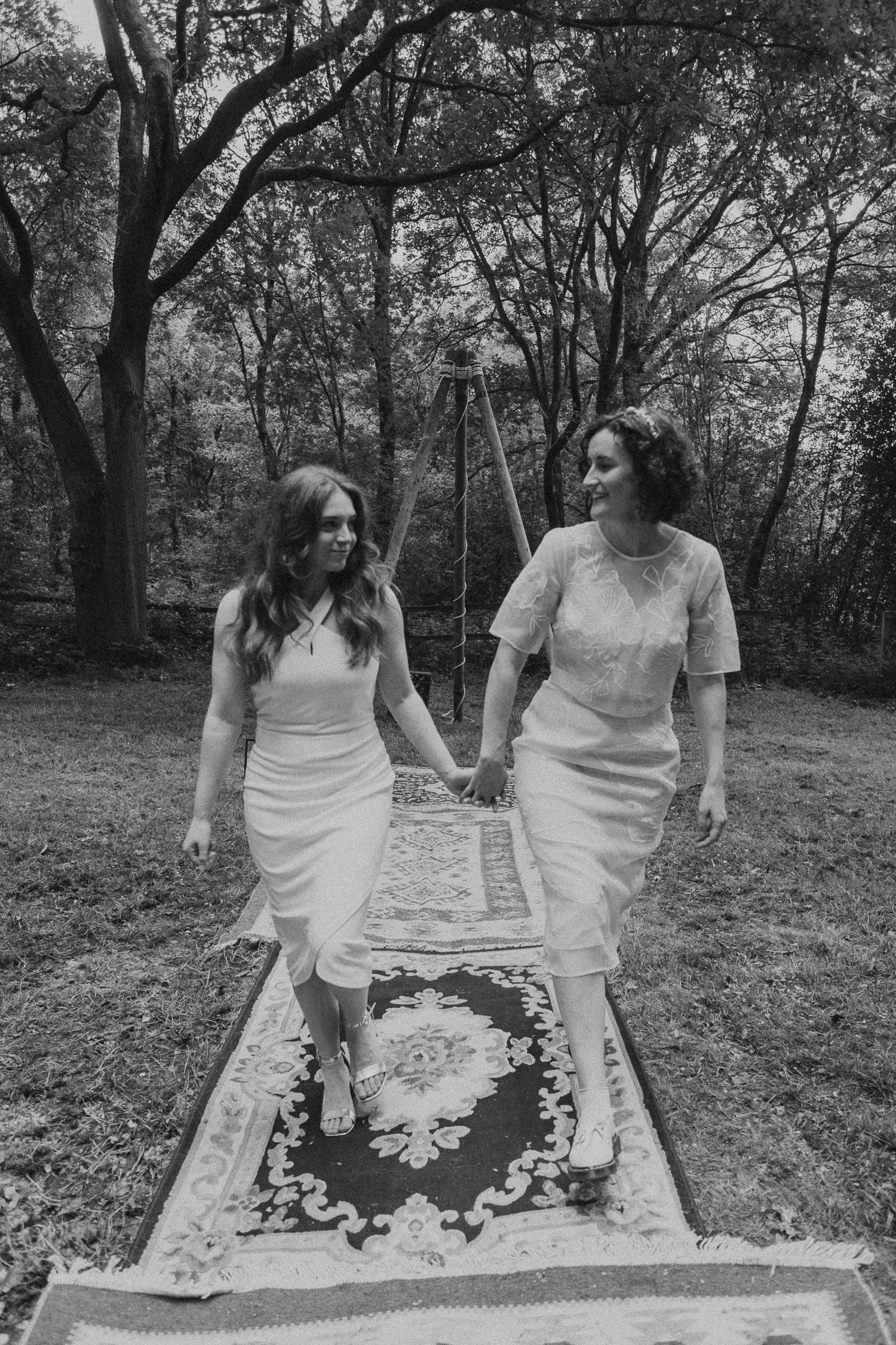 Two people in white dresses sit together on a patterned blanket in a wooded outdoor setting.