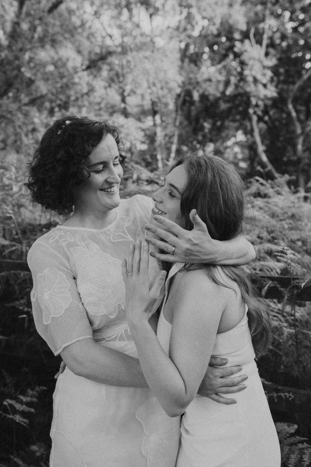 A tender black and white moment between two people in white dresses embracing outdoors among trees.