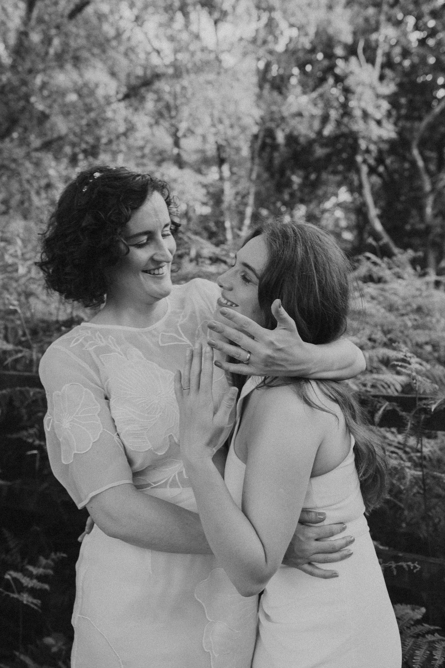 A tender black and white moment between two people in white dresses embracing outdoors among trees.