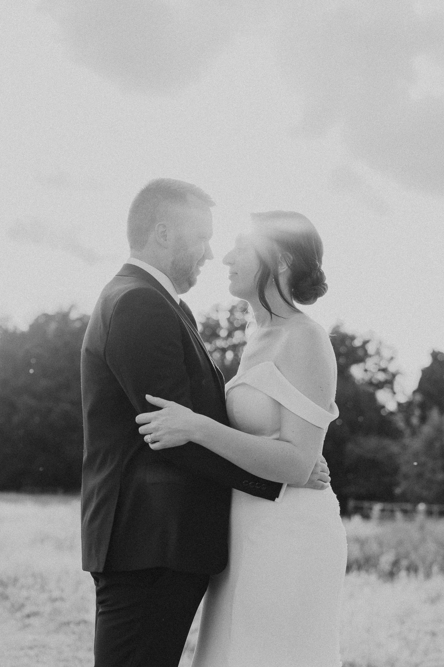 Romantic black and white photo of a couple in wedding attire embracing in a sunlit outdoor field.
