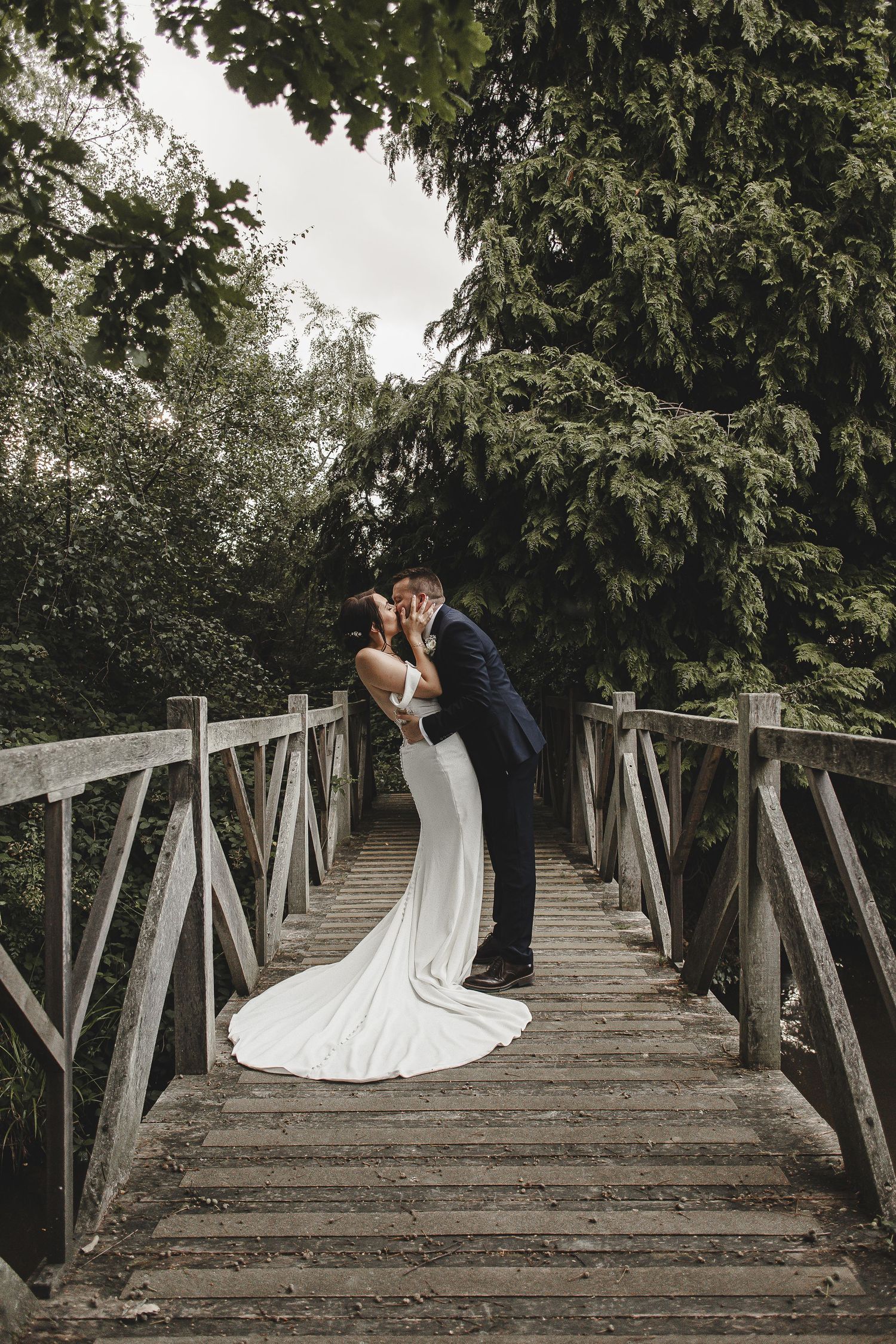 A couple shares a romantic moment on a wooden footbridge surrounded by lush green trees in their wedding attire.