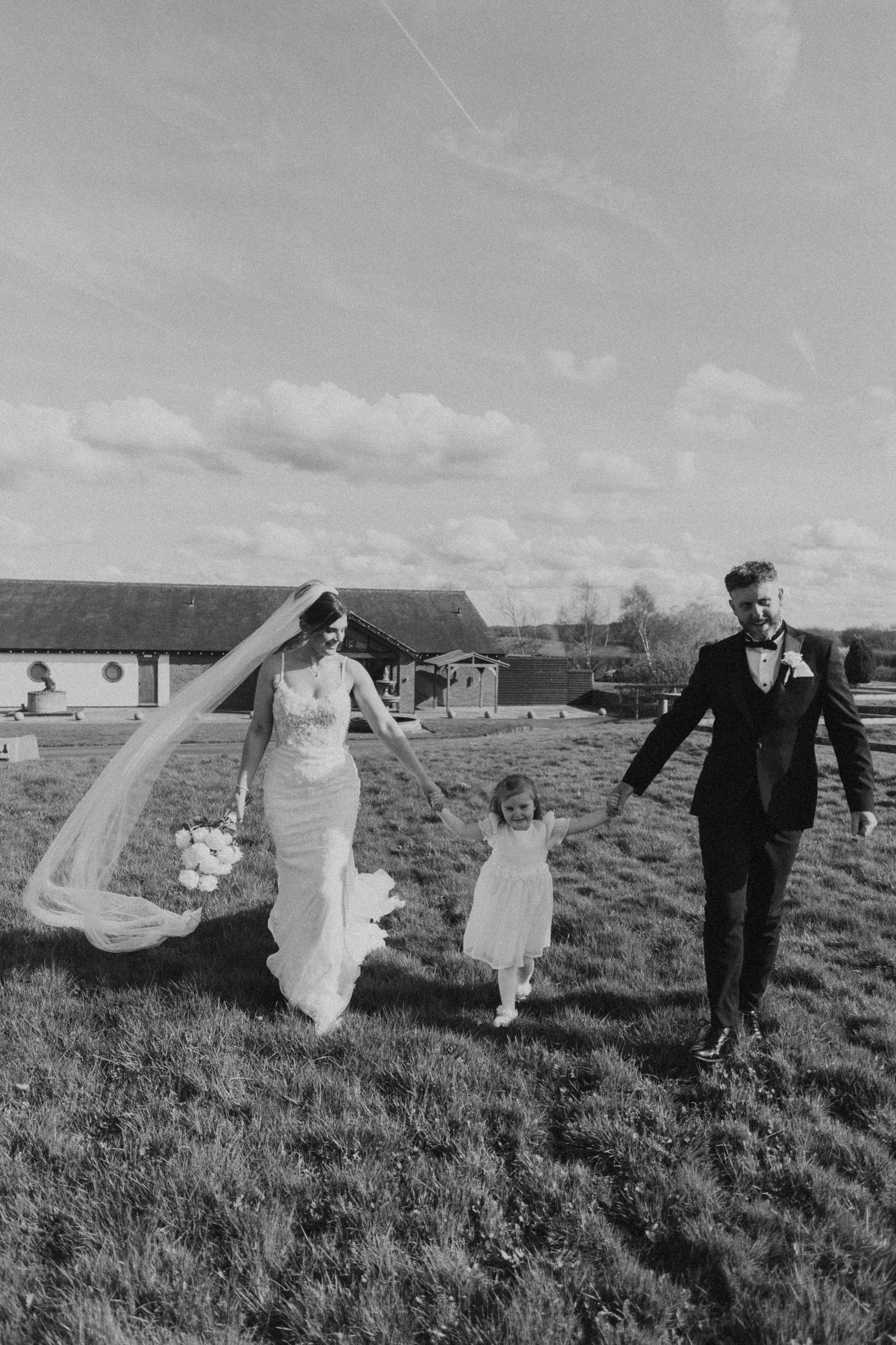 A wedding party runs playfully through a grassy field on an overcast day, with their formal attire flowing in the wind.
