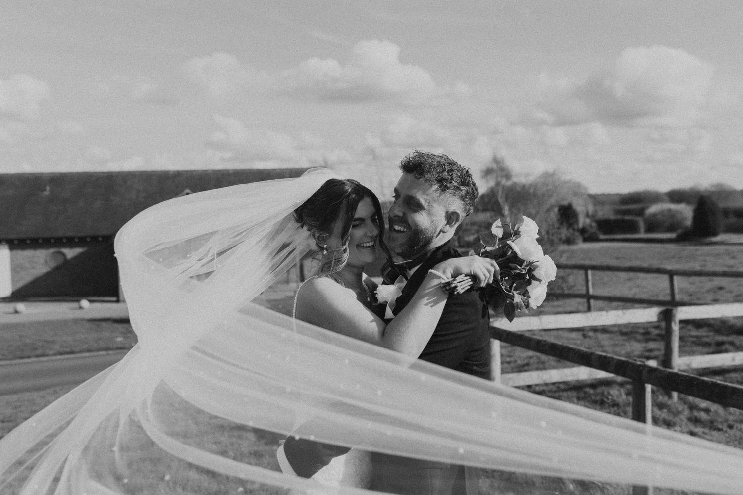 A long white wedding veil flows dramatically in the wind against a rural landscape backdrop.