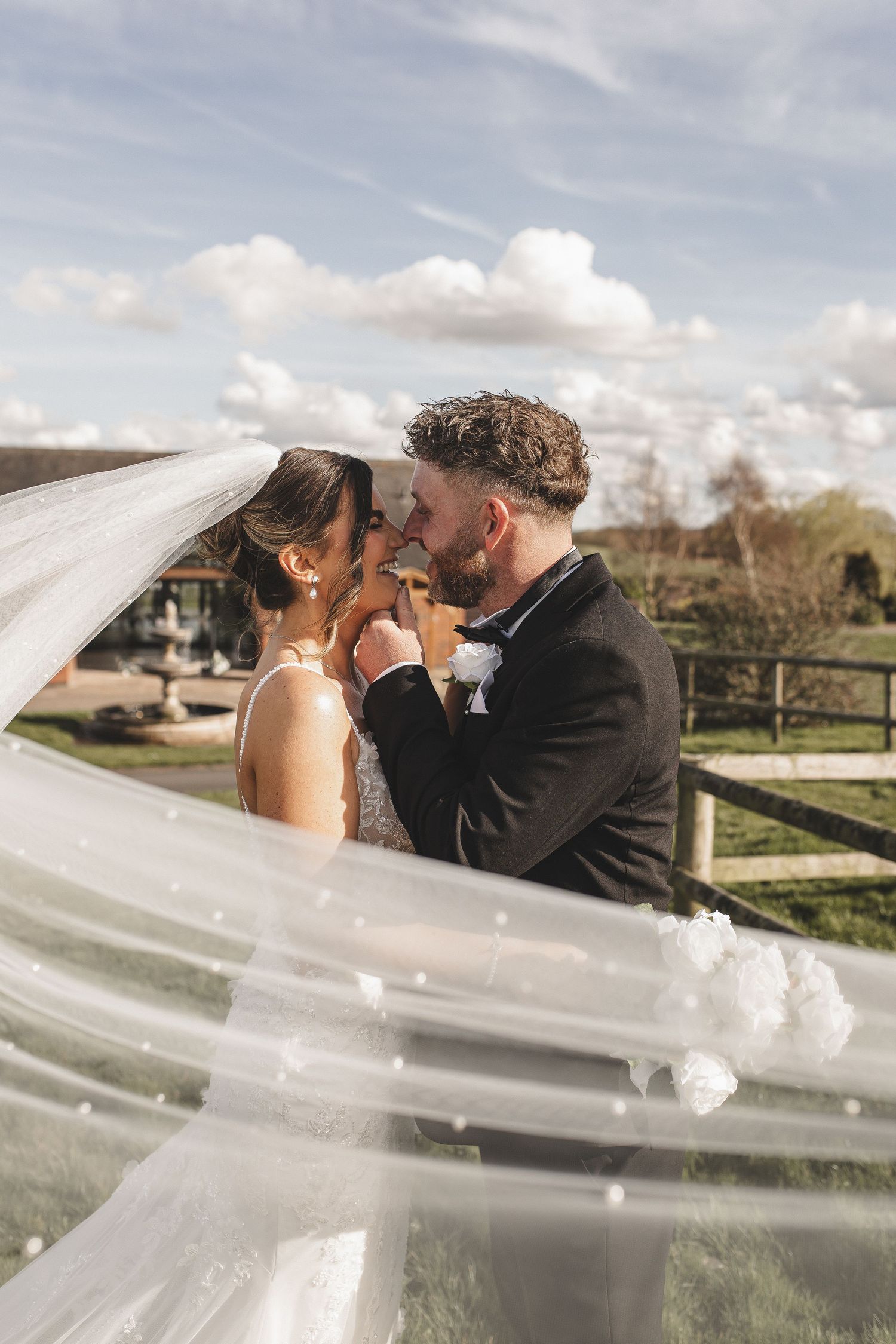 A romantic wedding moment captured through a flowing veil against a cloudy sky.