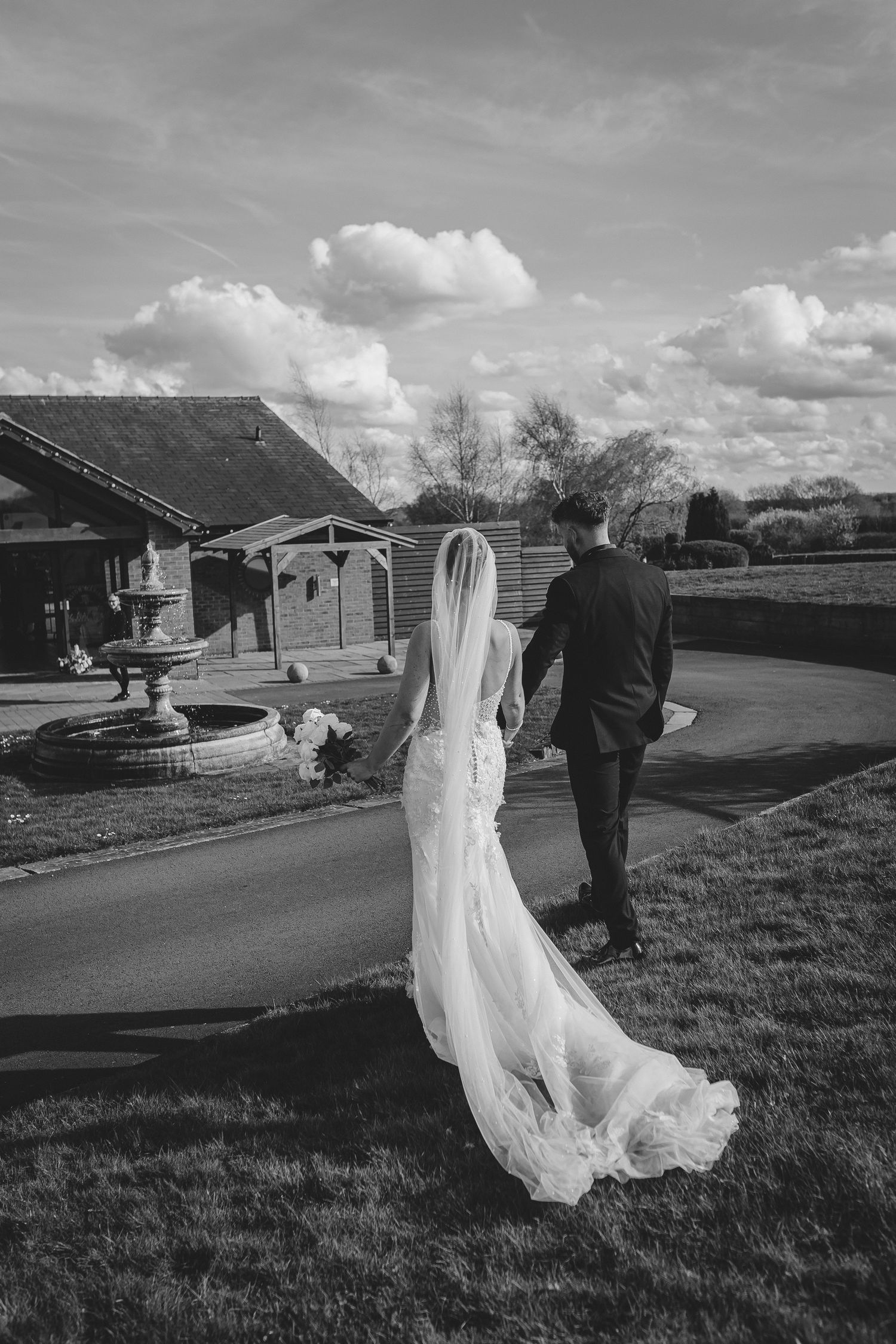 A couple walks along a path with the bride's dress and veil flowing dramatically in black and white.