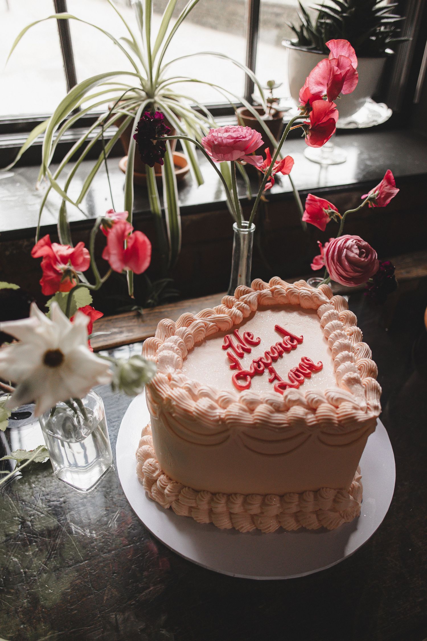 Heart-shaped pink frosted cake with decorative piping sits on a white plate near orchid flowers and a window.