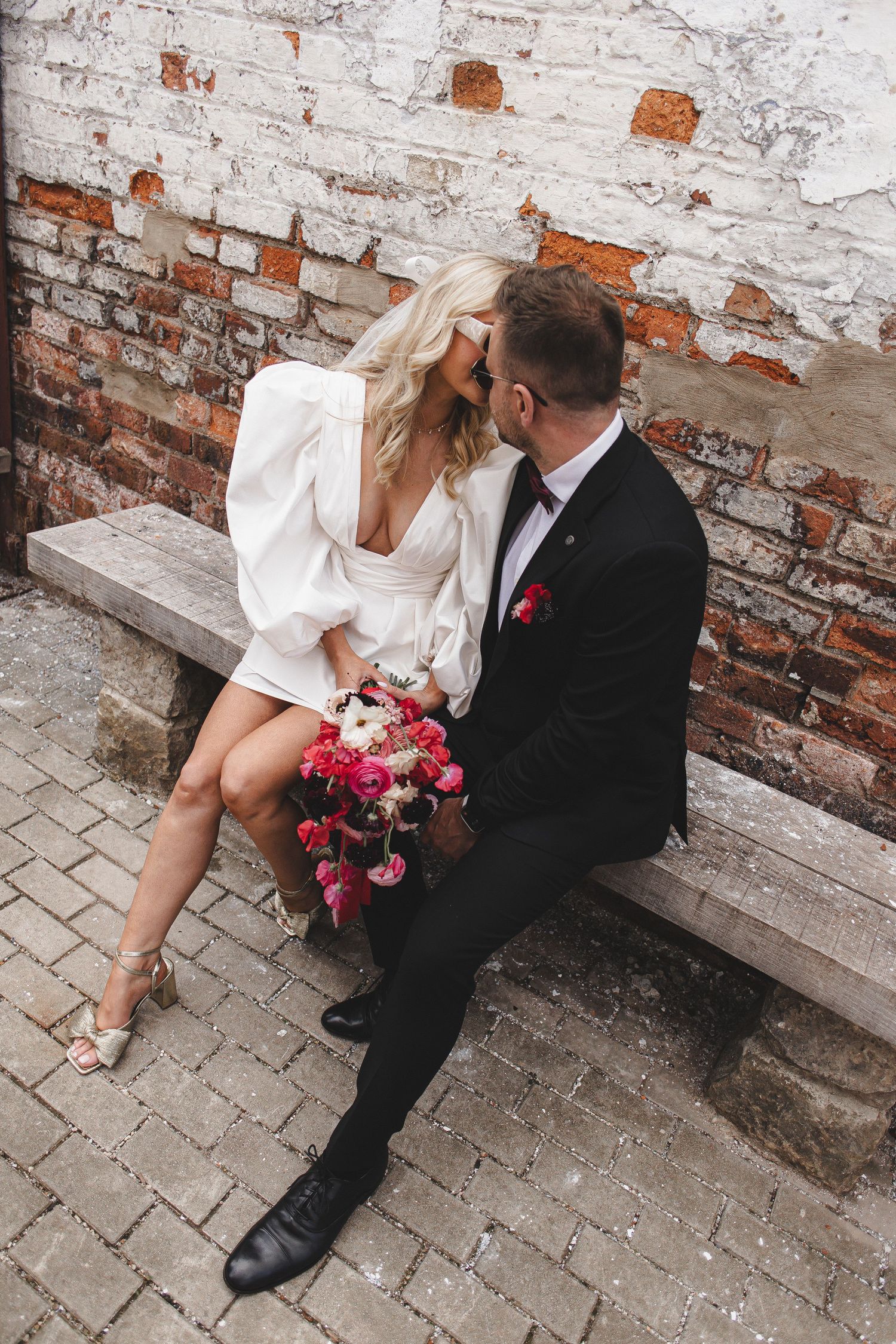 Newlyweds sit on stone bench against weathered brick wall with pink floral bouquet.