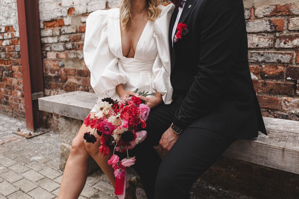 A couple in formal wedding attire embraces in front of a rustic brick wall with pink floral bouquet details.