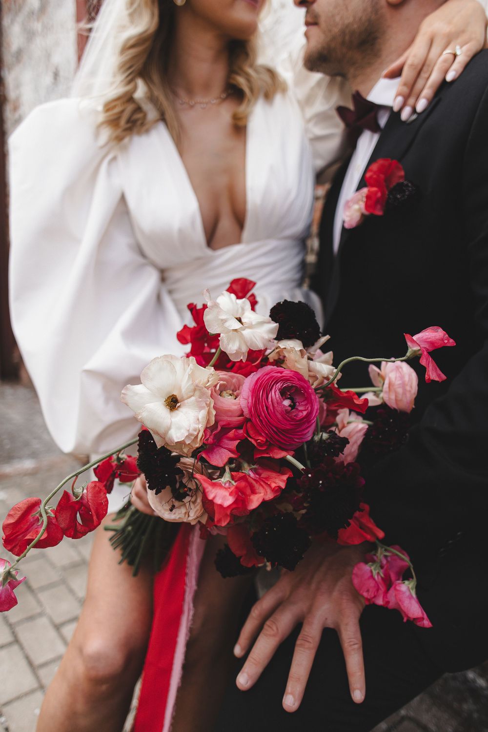 A vibrant red and pink wedding bouquet held between a couple in formal attire with white dress and black suit.