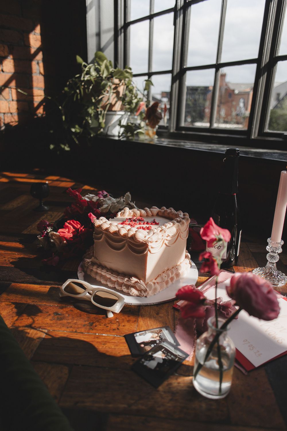 A white wedding cake with pink flowers sits on a rustic wooden table near windows in moody lighting.