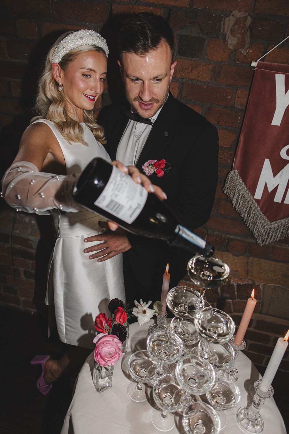 Couple celebrates near vintage silver tea set with pink roses on rustic brick backdrop.