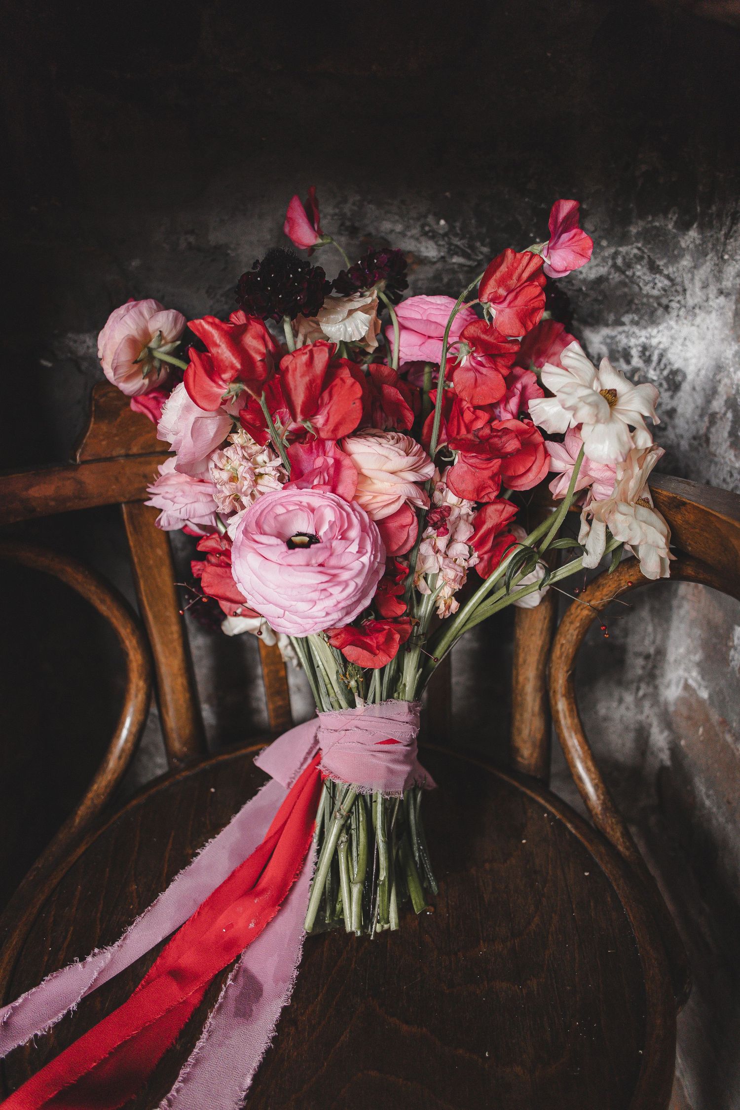 A romantic wedding bouquet with pink ranunculus, red flowers and trailing ribbons sits on a vintage wooden chair.