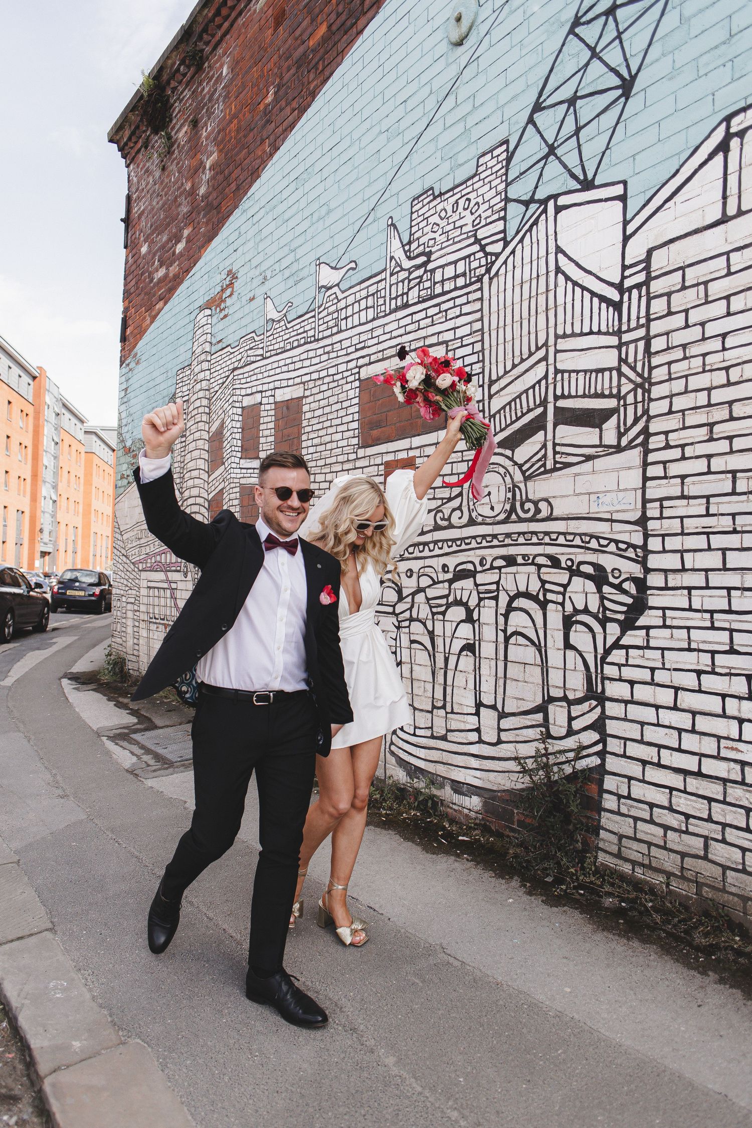 Joyful couple celebrates in front of a large architectural mural on white brick wall in urban setting.