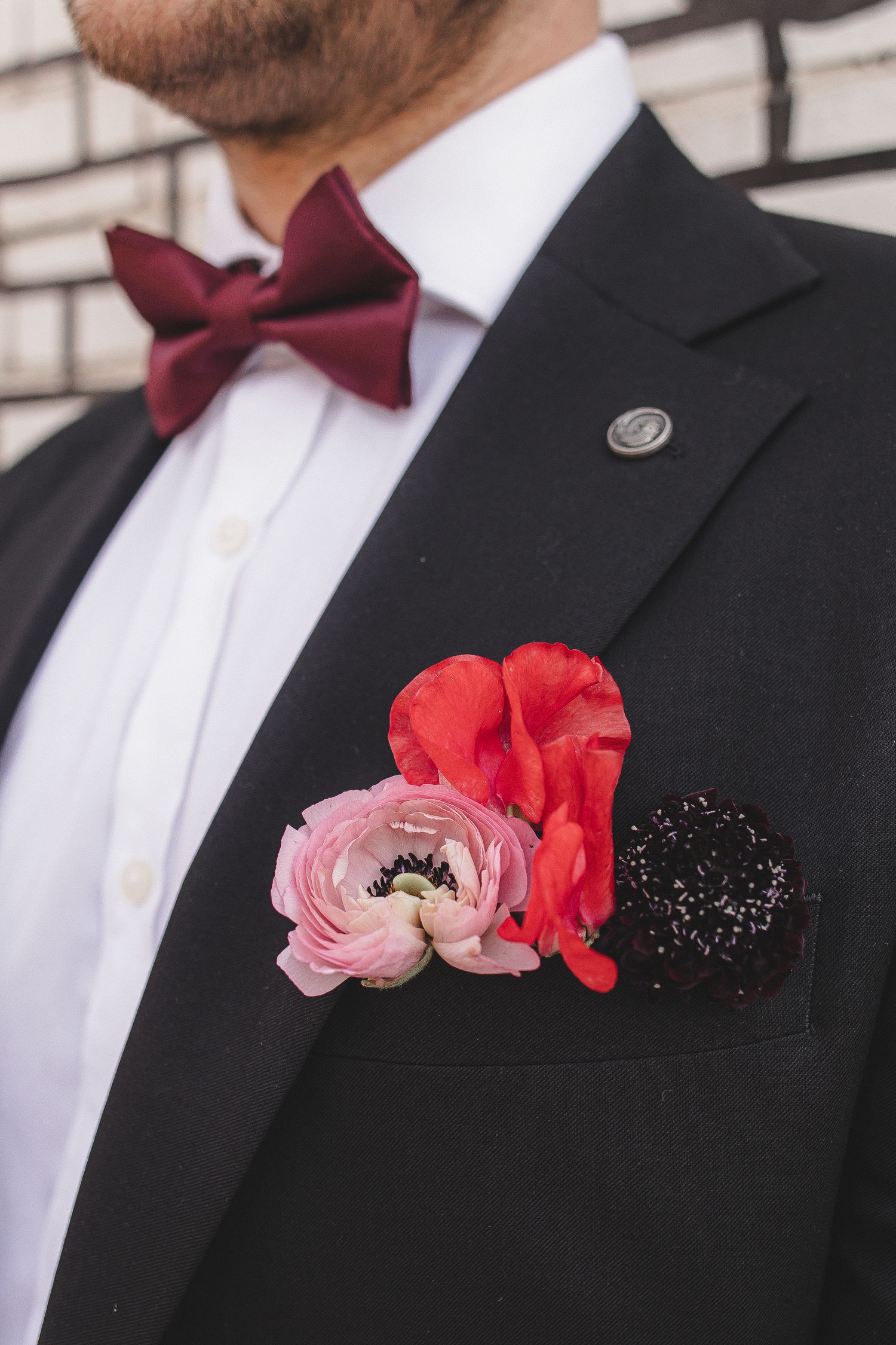 Close-up of formal black suit with burgundy bow tie and pink and red floral boutonniere against white brick wall.