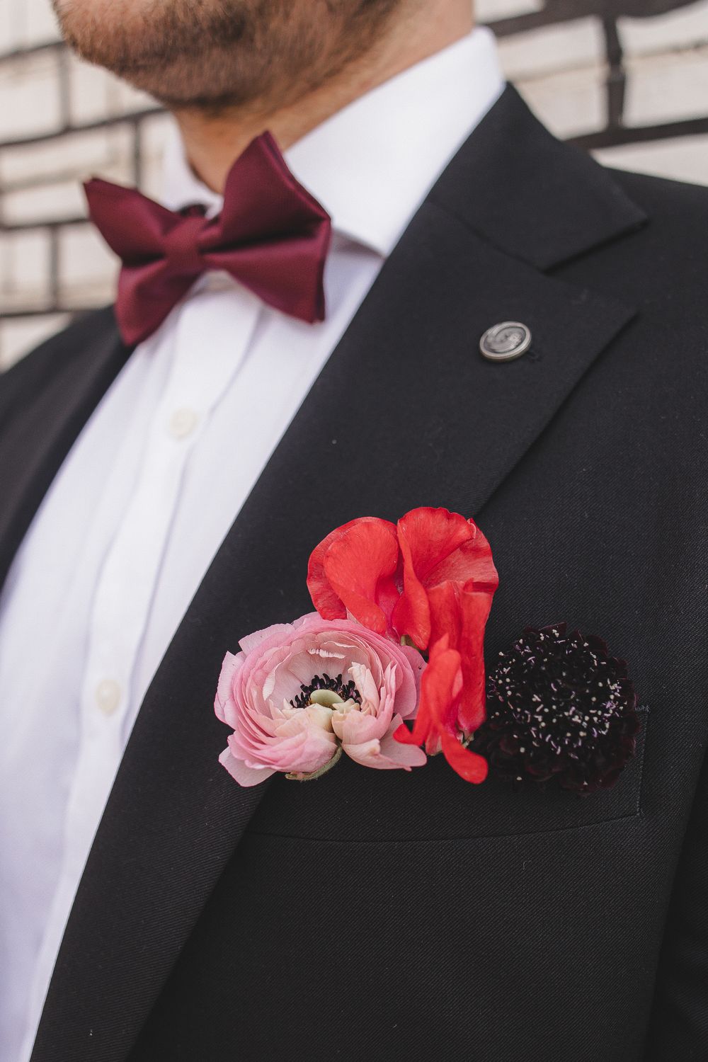 Close-up of formal black suit with burgundy bow tie and pink and red floral boutonniere against white brick wall.
