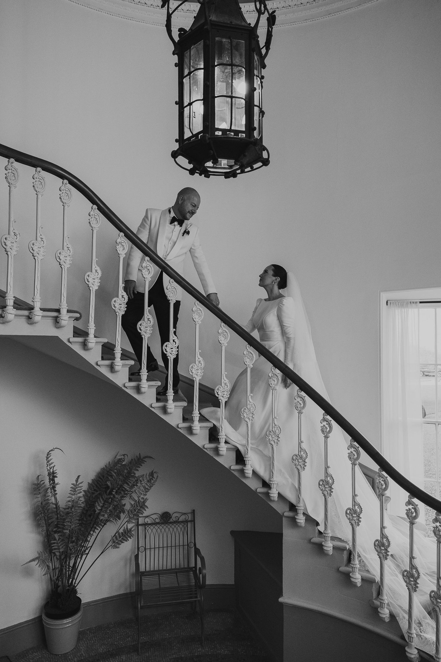 A black and white photograph of a couple on an elegant curved staircase with ornate railings and a hanging lantern above.