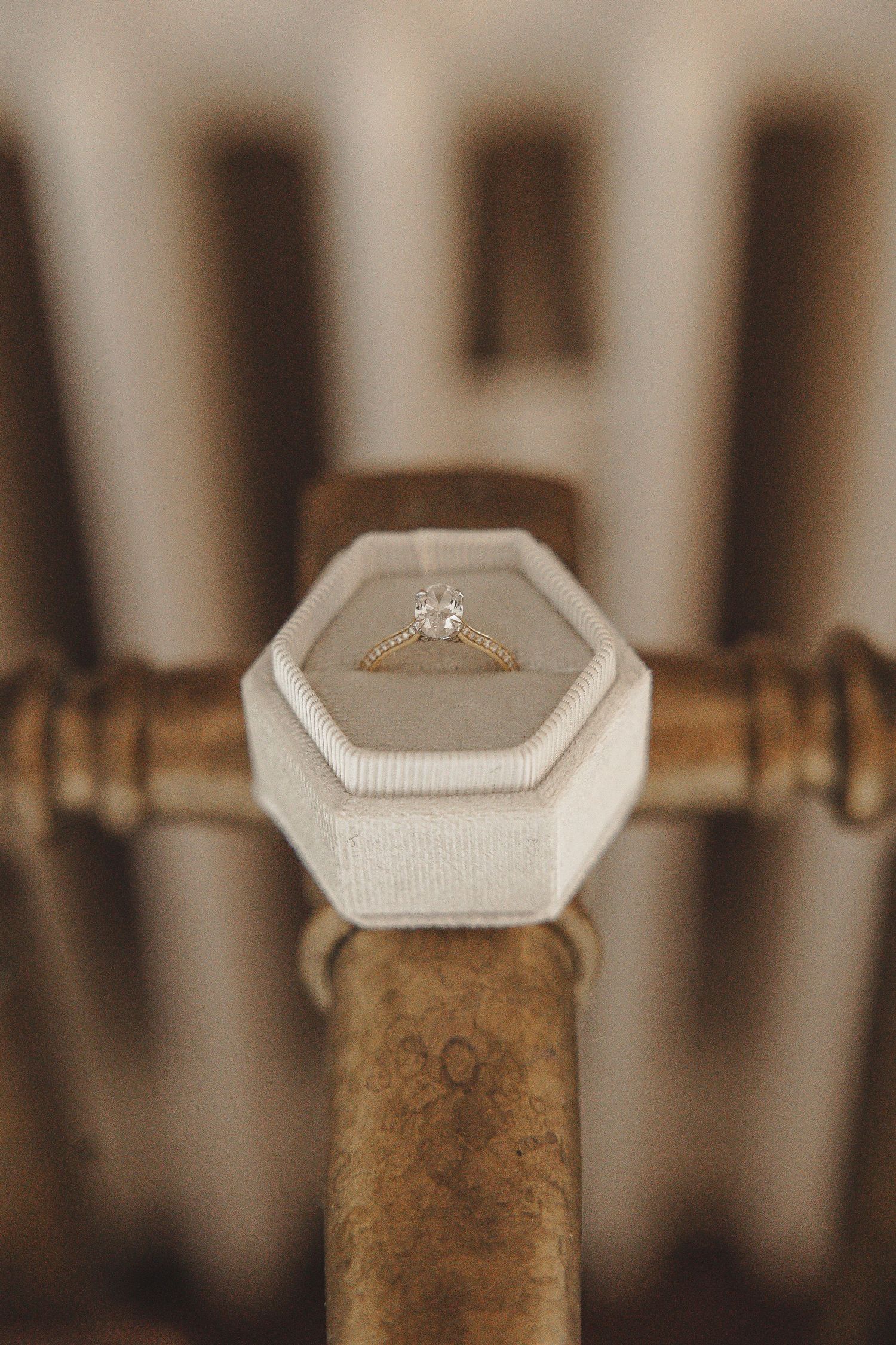Diamond engagement ring in white hexagonal box balanced on vintage brass railing with blurred background.