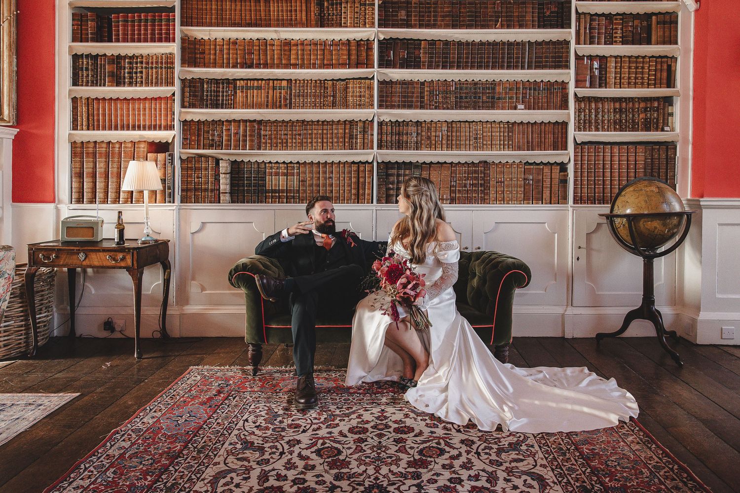 A couple shares a romantic moment in a grand library with red walls and vintage books on wooden shelves.