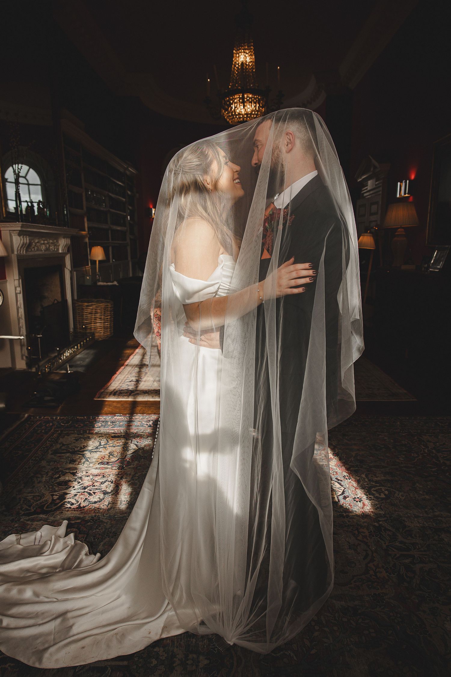 A romantic wedding couple embraces under a flowing veil in a dimly lit venue with elegant chandeliers overhead.
