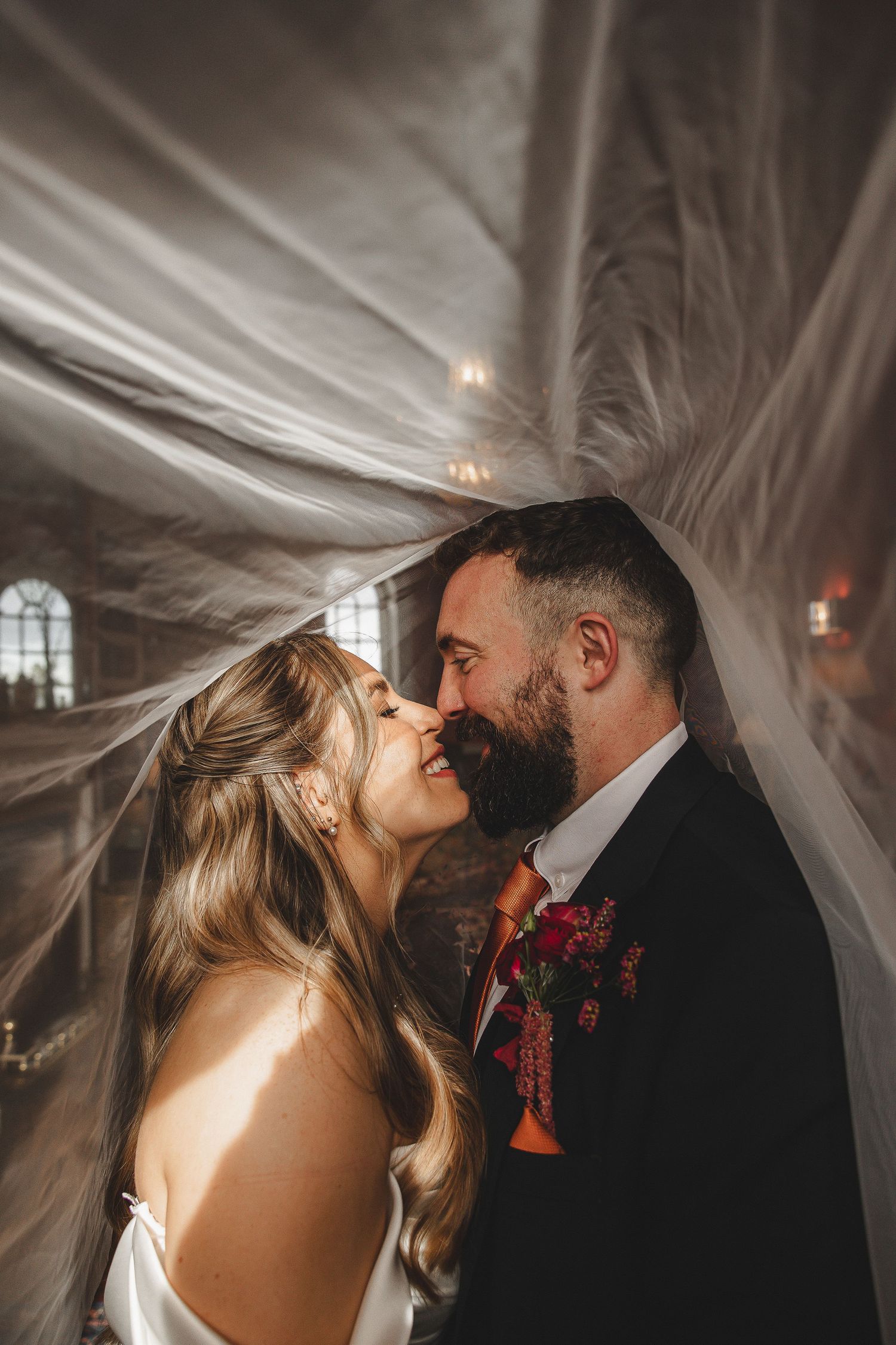 Romantic wedding couple shares an intimate moment under flowing veil with dramatic lighting in church setting.