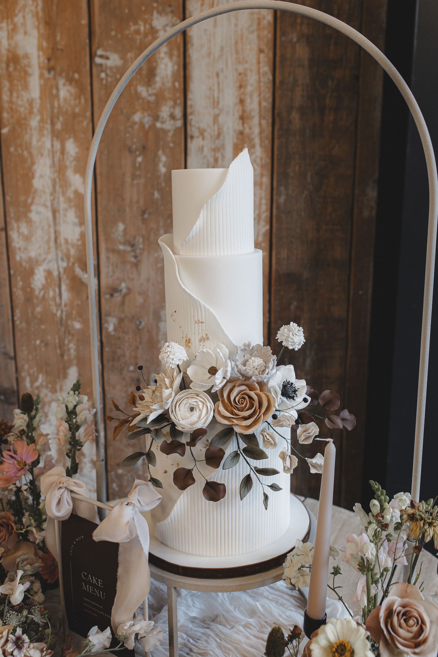 Elegant white wedding cake decorated with delicate rose gold and white flowers on rustic wooden backdrop.