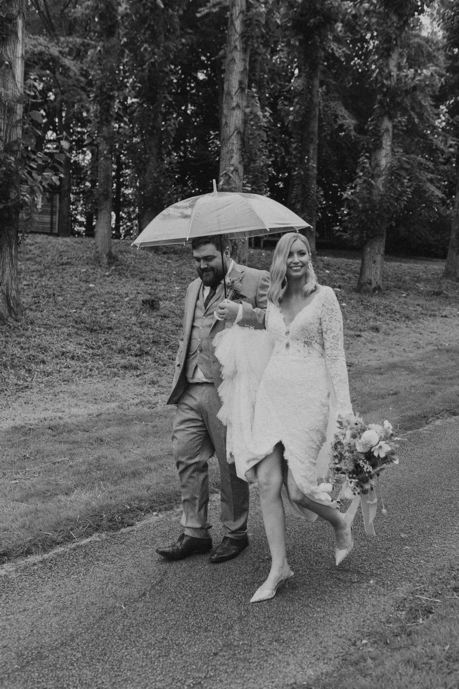 A couple in wedding attire shares a playful moment under an umbrella in a wooded outdoor setting.