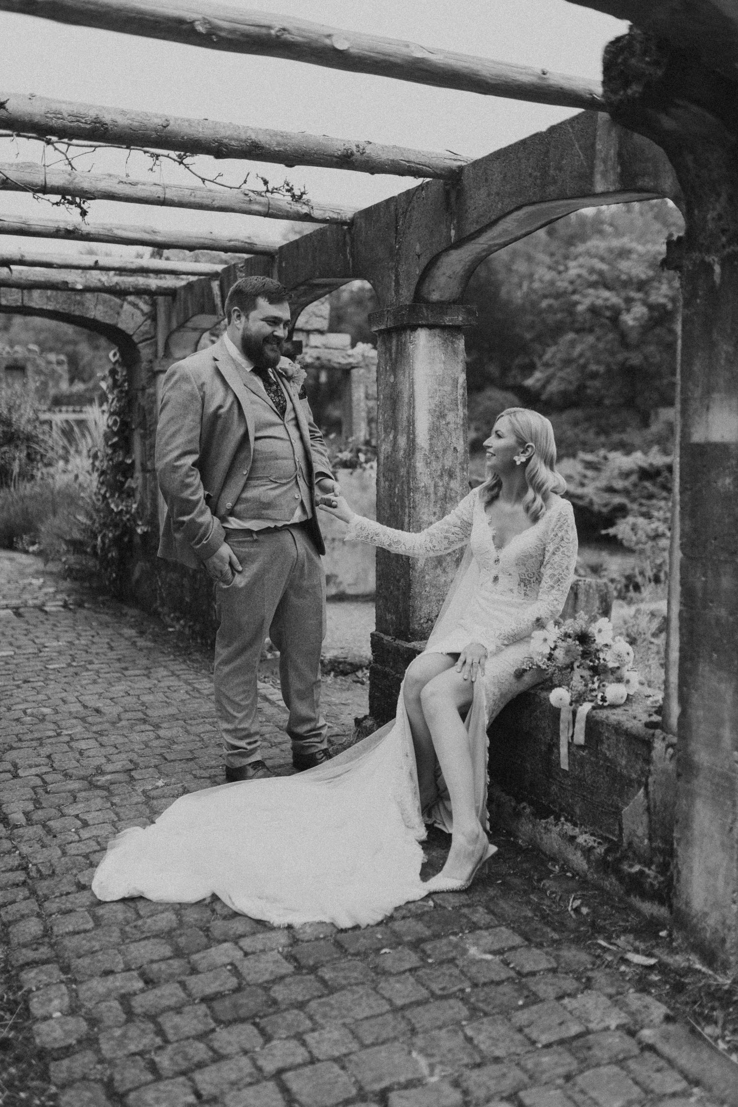 Black and white wedding photos show couple posing romantically under rustic stone archway on cobblestone path.
