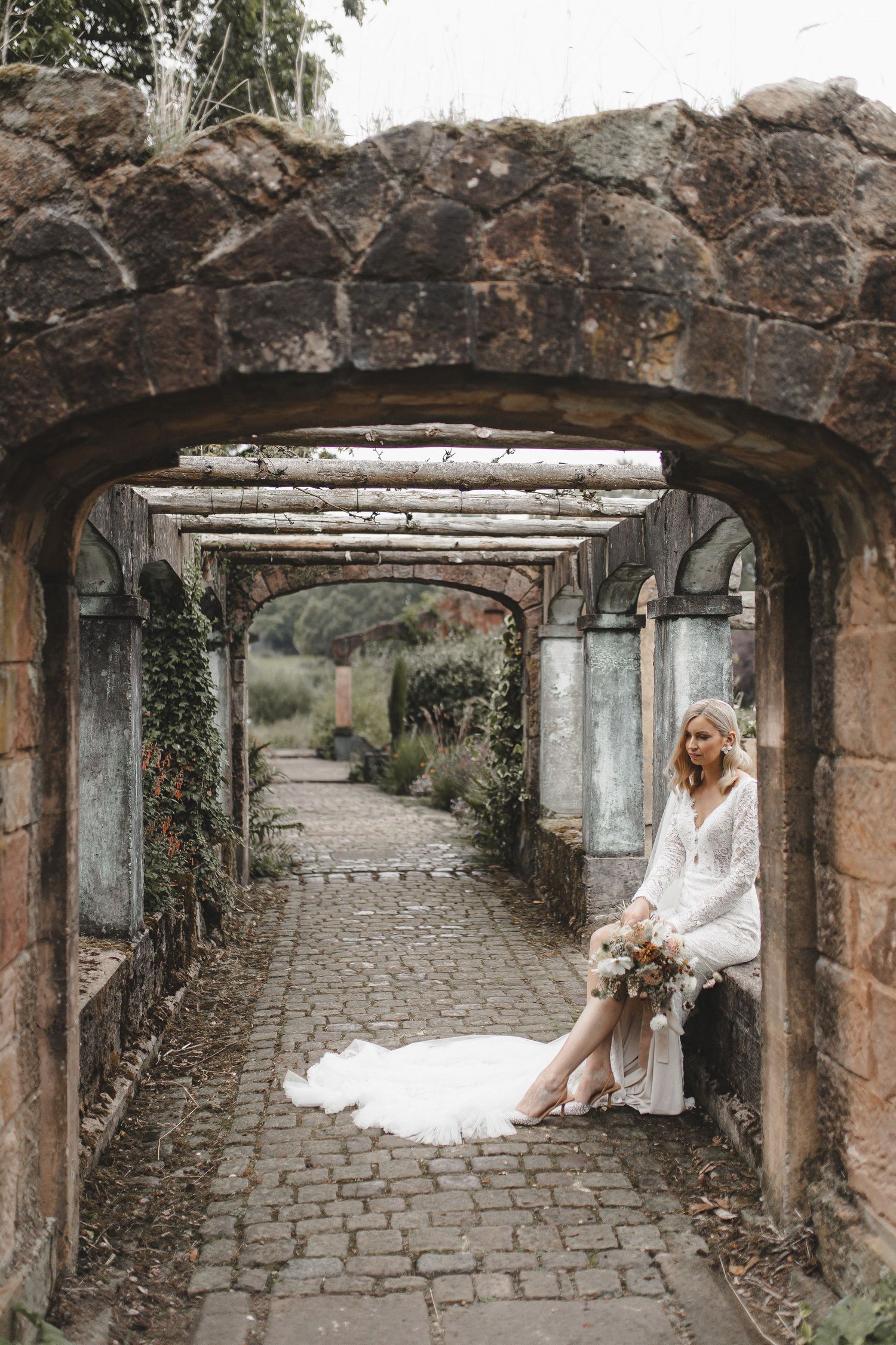 A stone archway covered in ivy leads down a cobblestone path where a white wedding dress creates a romantic scene.