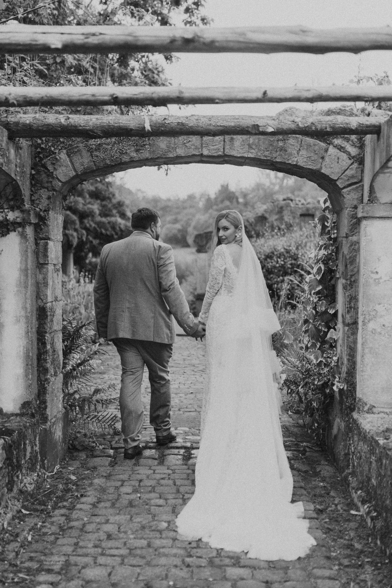 A romantic black and white wedding photo showing a couple walking through a stone archway in a garden setting.