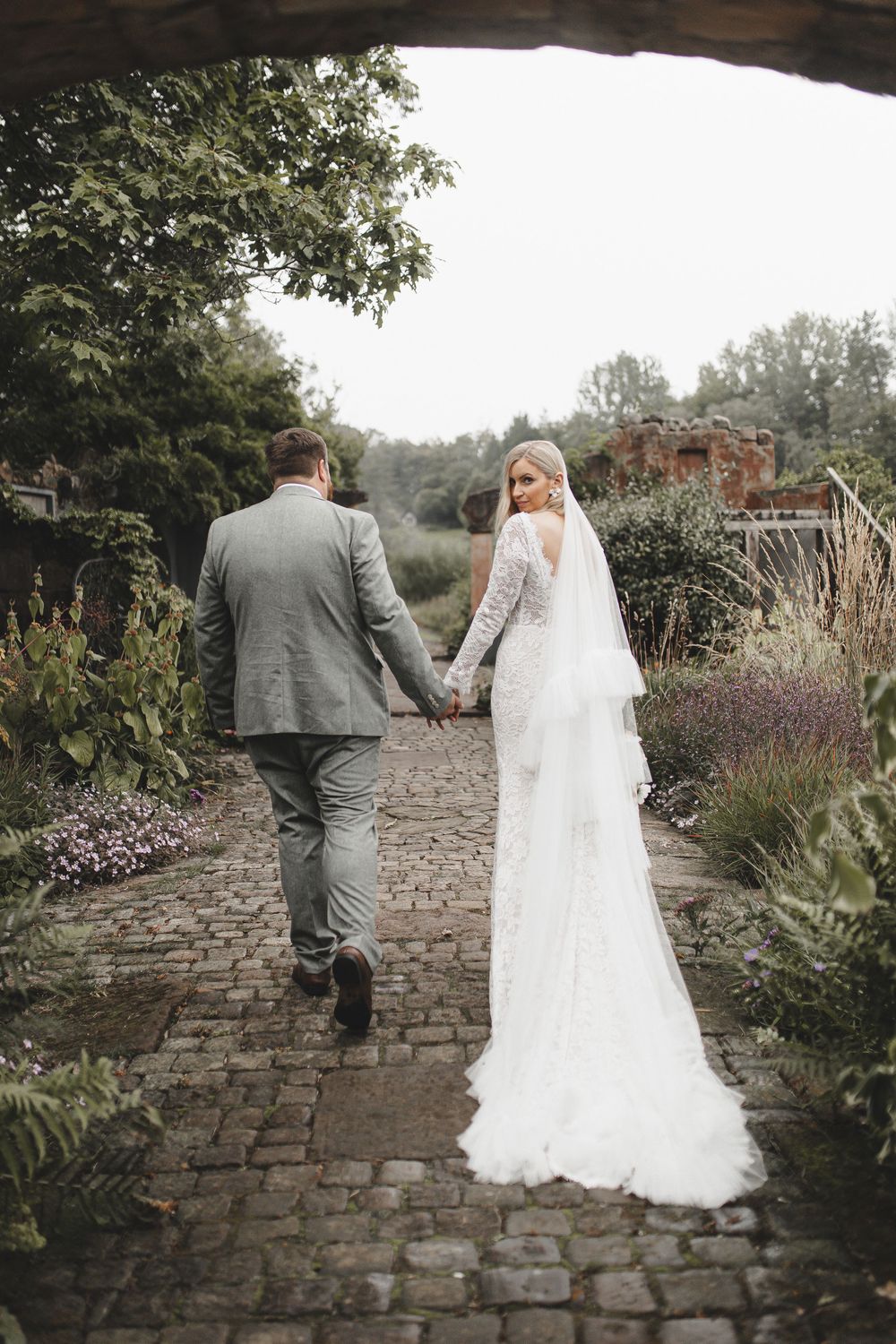 A couple in wedding attire walks along a rustic stone path bordered by lush garden flowers and greenery.