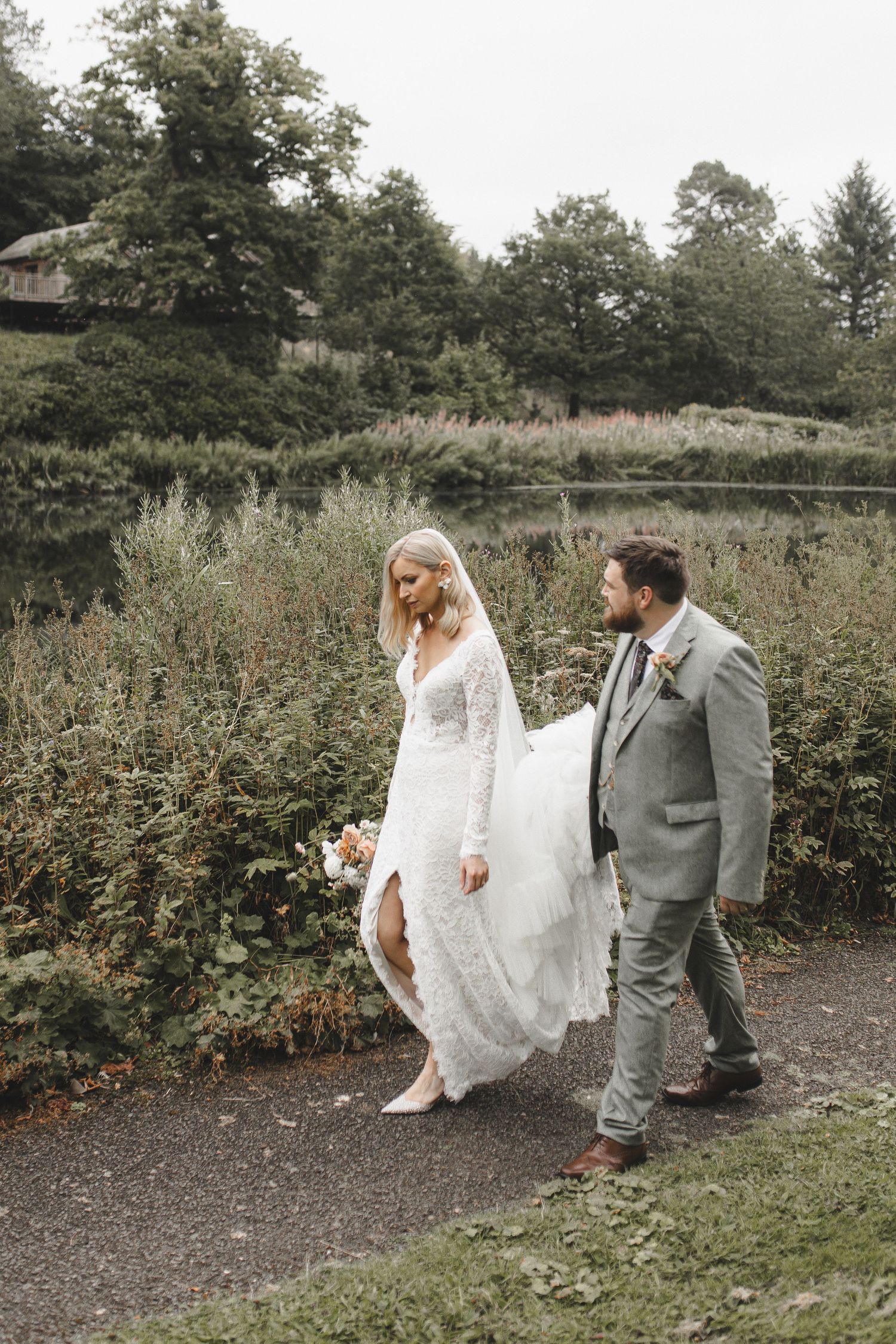 A wedding couple walking along a garden path surrounded by lush greenery and wildflowers.