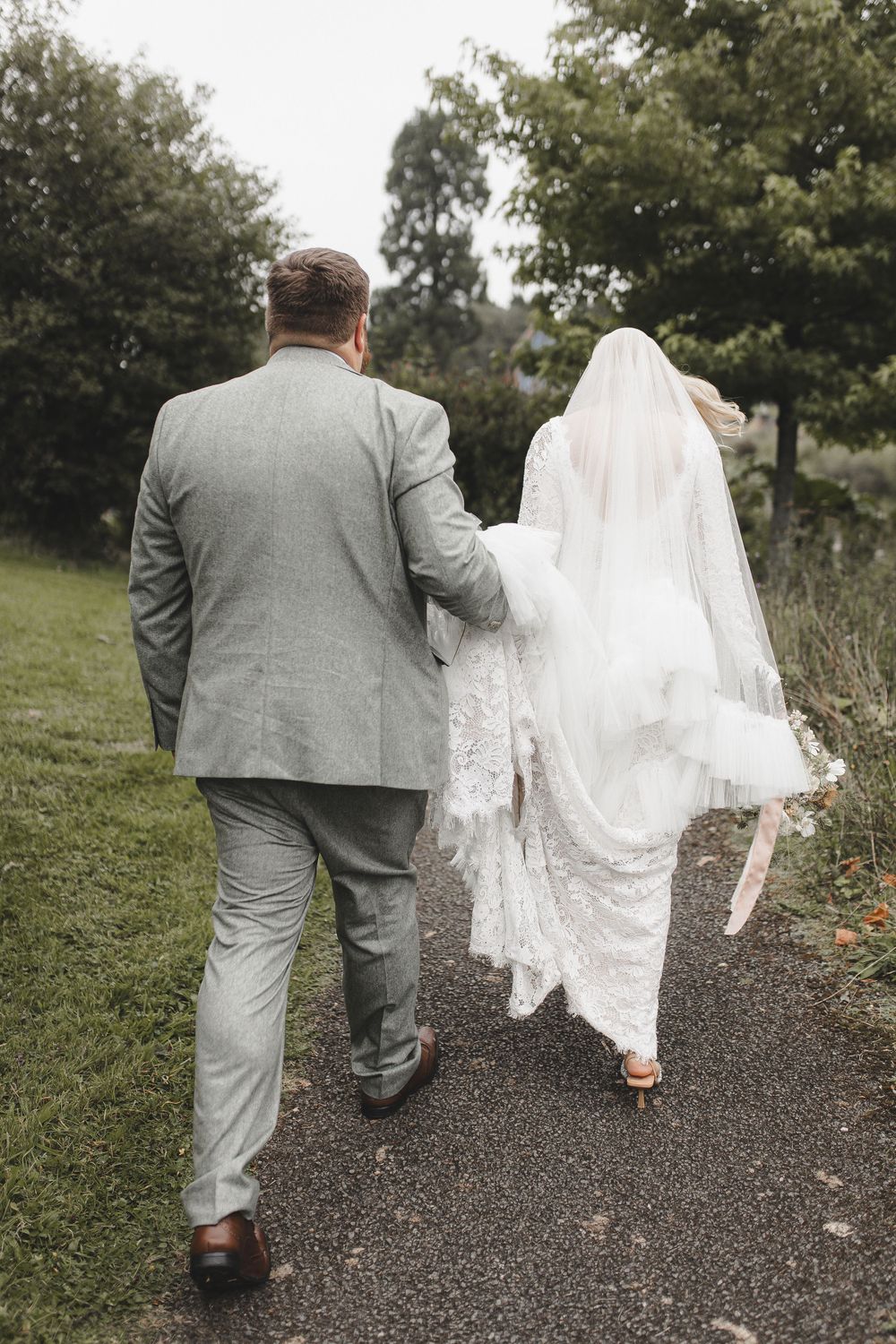 A couple walking down a gravel path, with the bride's lace veil flowing behind her and groom in a gray suit.
