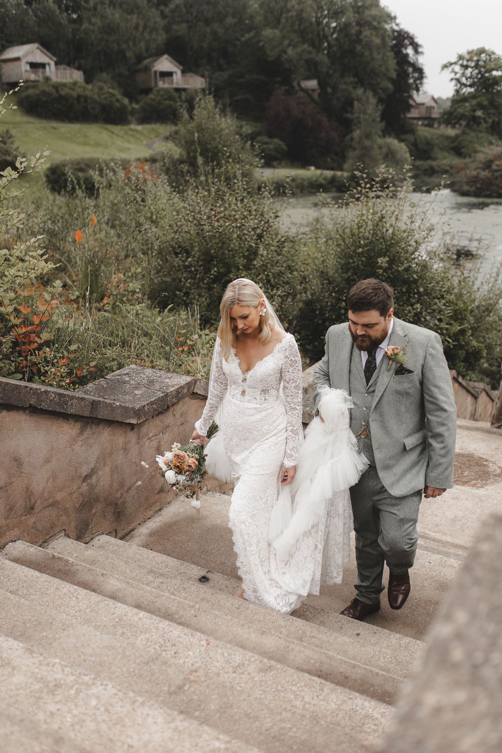 A couple descends stone steps at a waterfront venue, with rustic cabins visible on the hillside behind them.