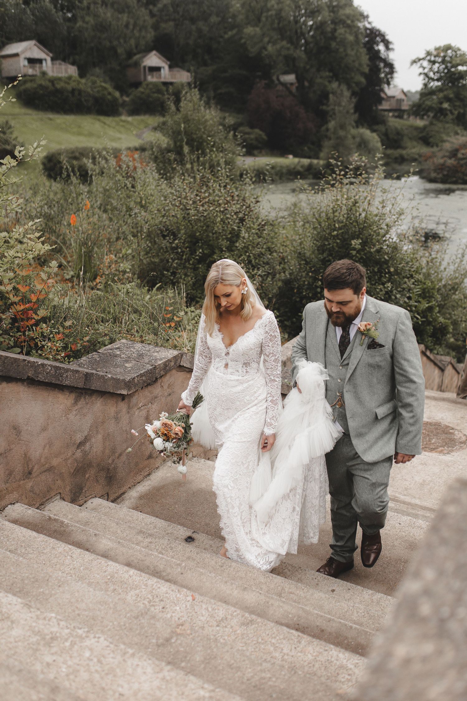 A couple descends stone steps at a waterfront venue, with rustic cabins visible on the hillside behind them.