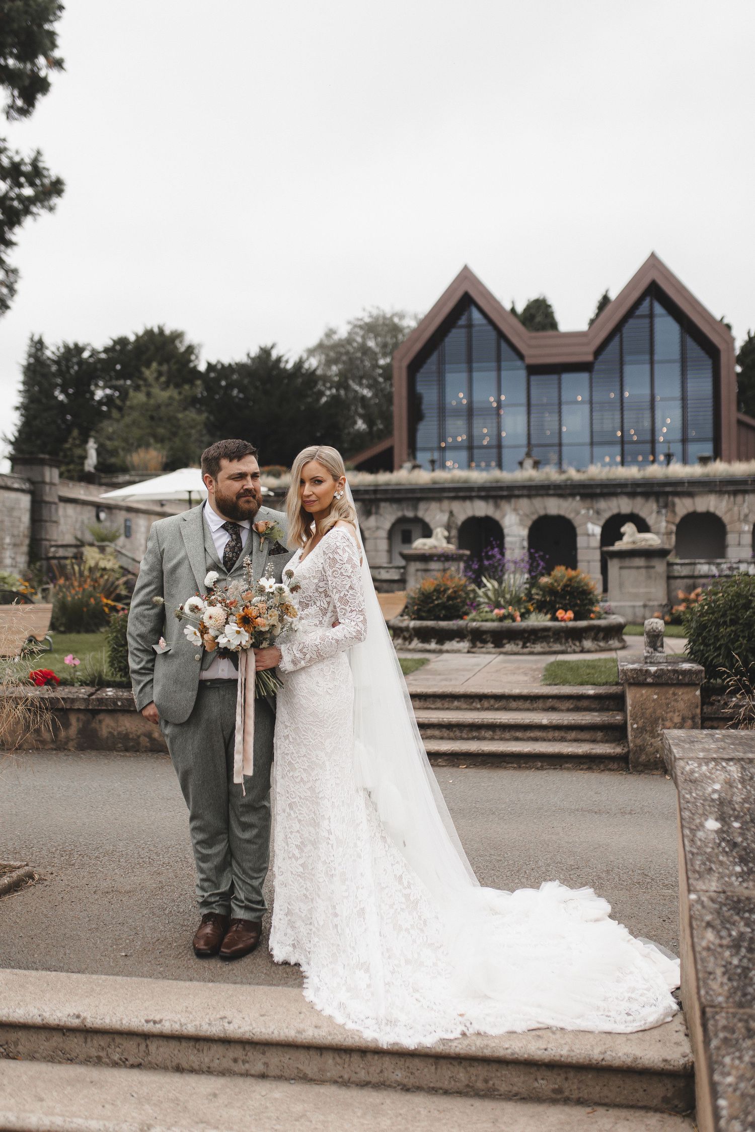 A modern chapel with distinctive peaked glass windows serves as backdrop for an elegant outdoor wedding portrait.