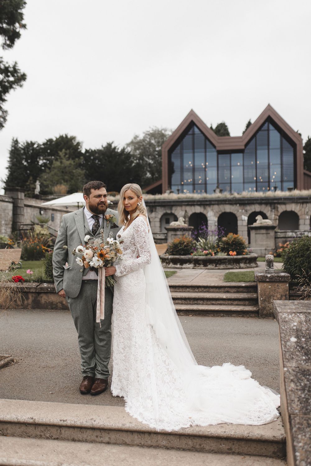 A modern chapel with distinctive peaked glass windows serves as backdrop for an elegant outdoor wedding portrait.