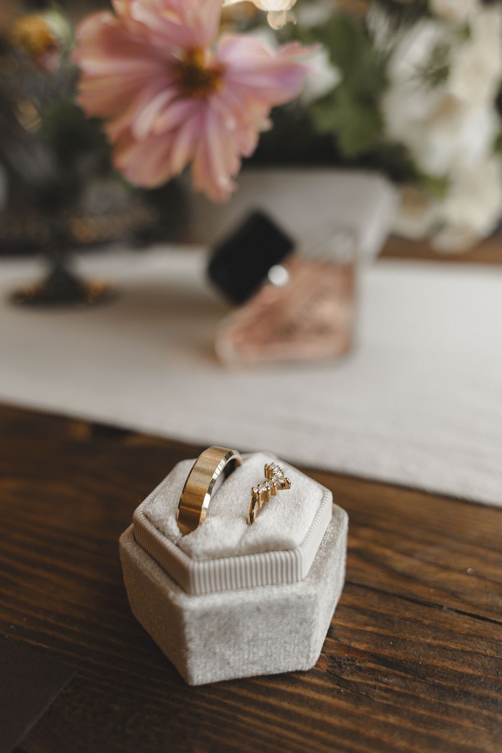 A delicate white hexagonal ring box with gold accents sits on a dark wooden surface with pink flowers in the background.